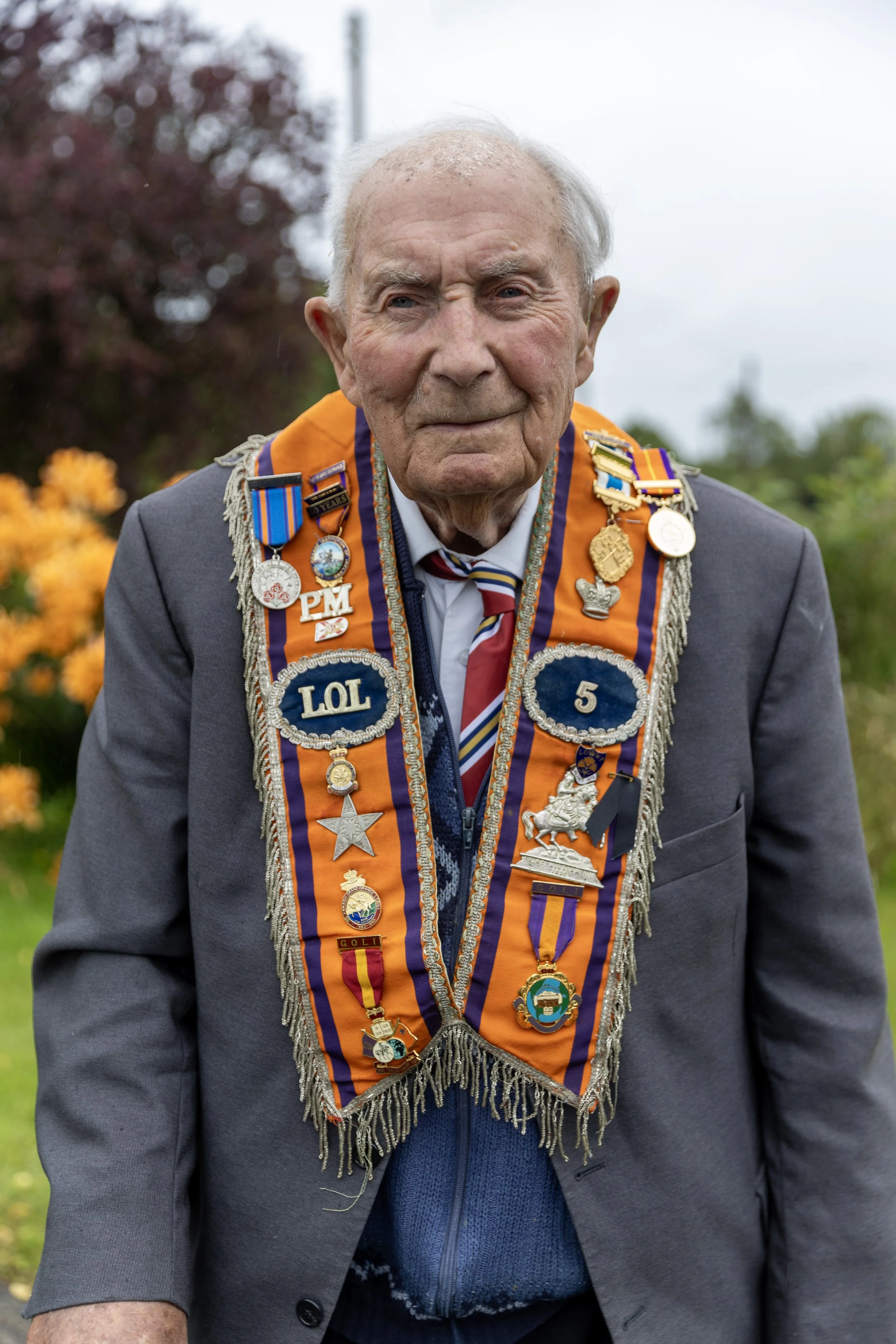 An elderly man dressed in a suit with a sash adorned with medals and badges, standing outdoors in front of colorful flowers and trees.