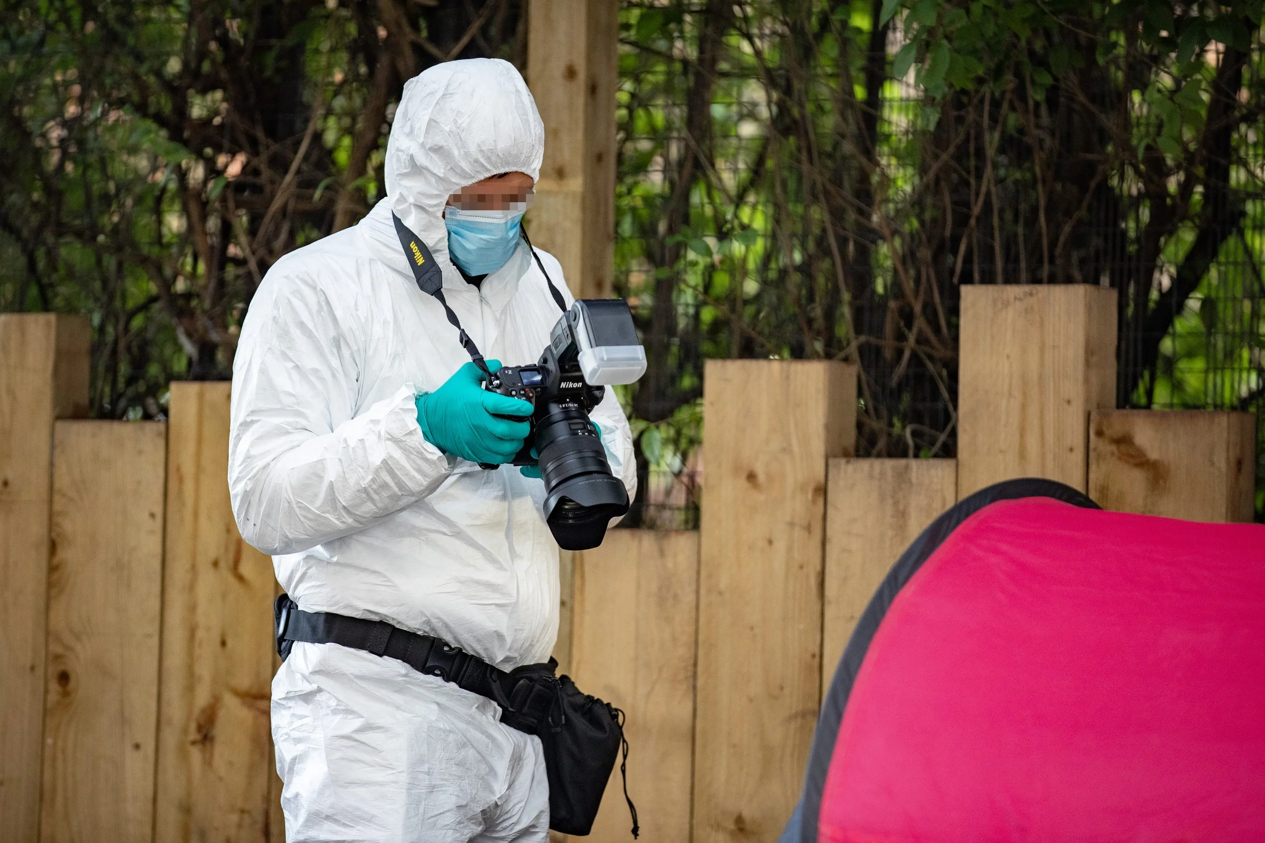 A person dressed in white protective suit, face mask, and teal gloves holding a professional camera, in an outdoor setting with a wooden fence and green foliage background.