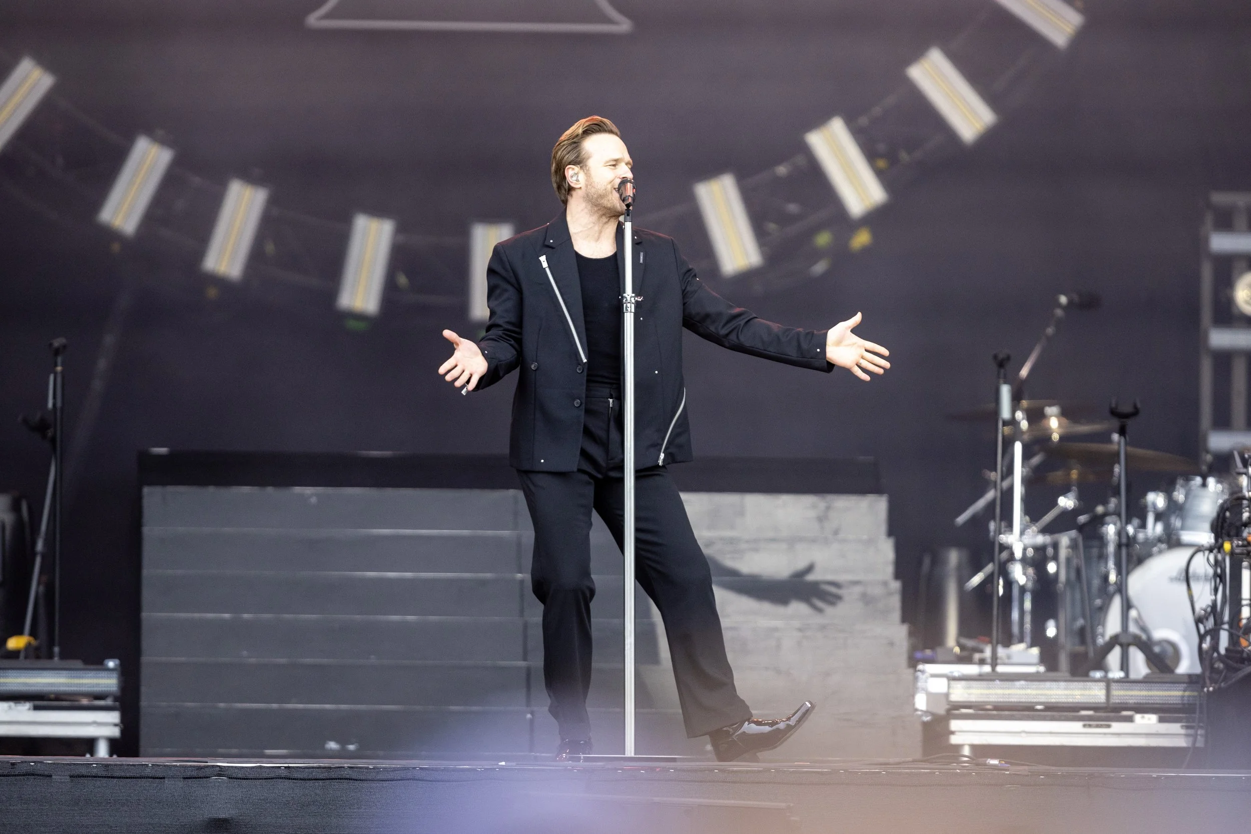 A male singer performing on stage with a microphone, wearing a black suit and gesturing with his arms open, with musical instruments in the background.