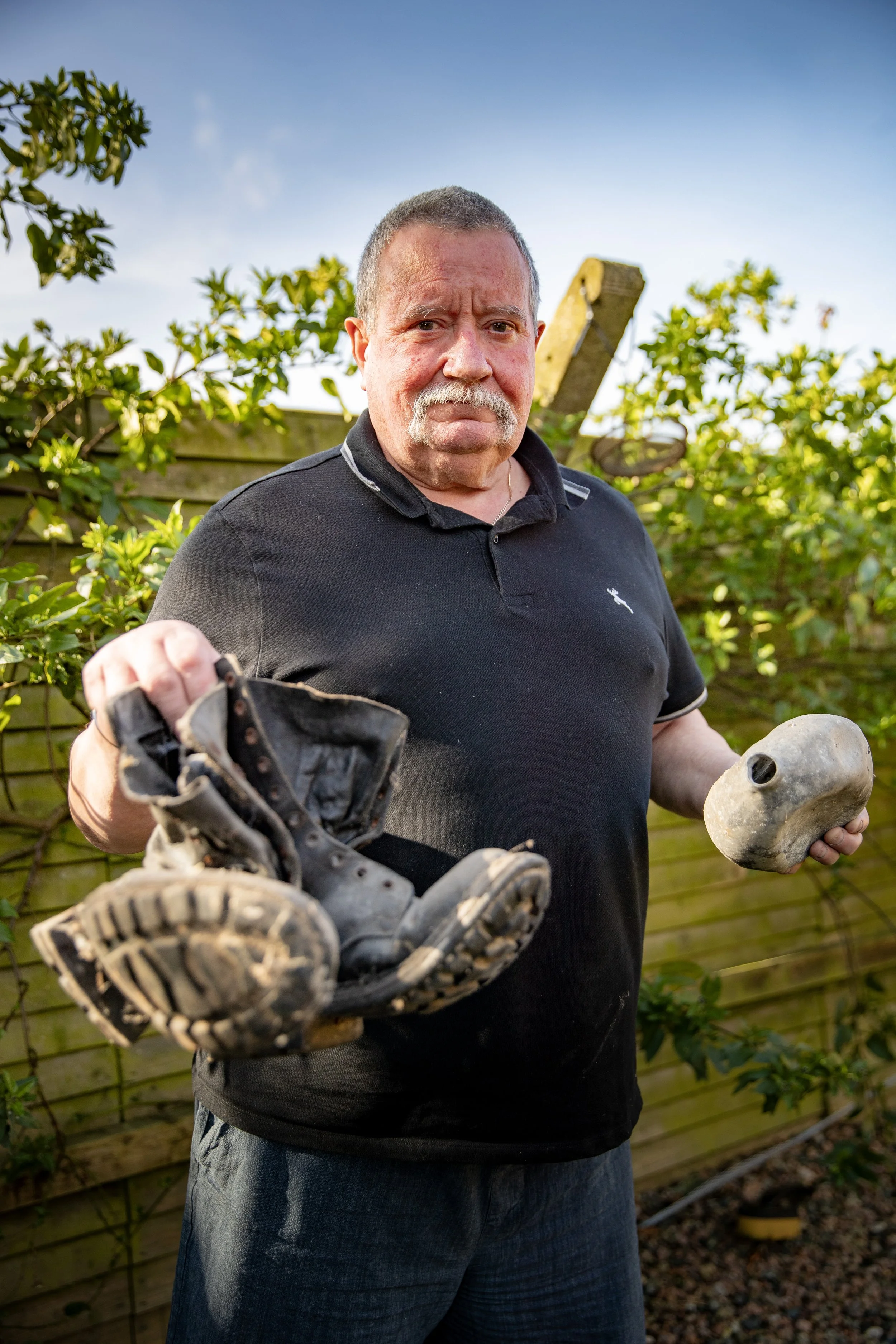 An older man with a mustache and short gray hair holding a worn-out shoe and a stone in a backyard garden.