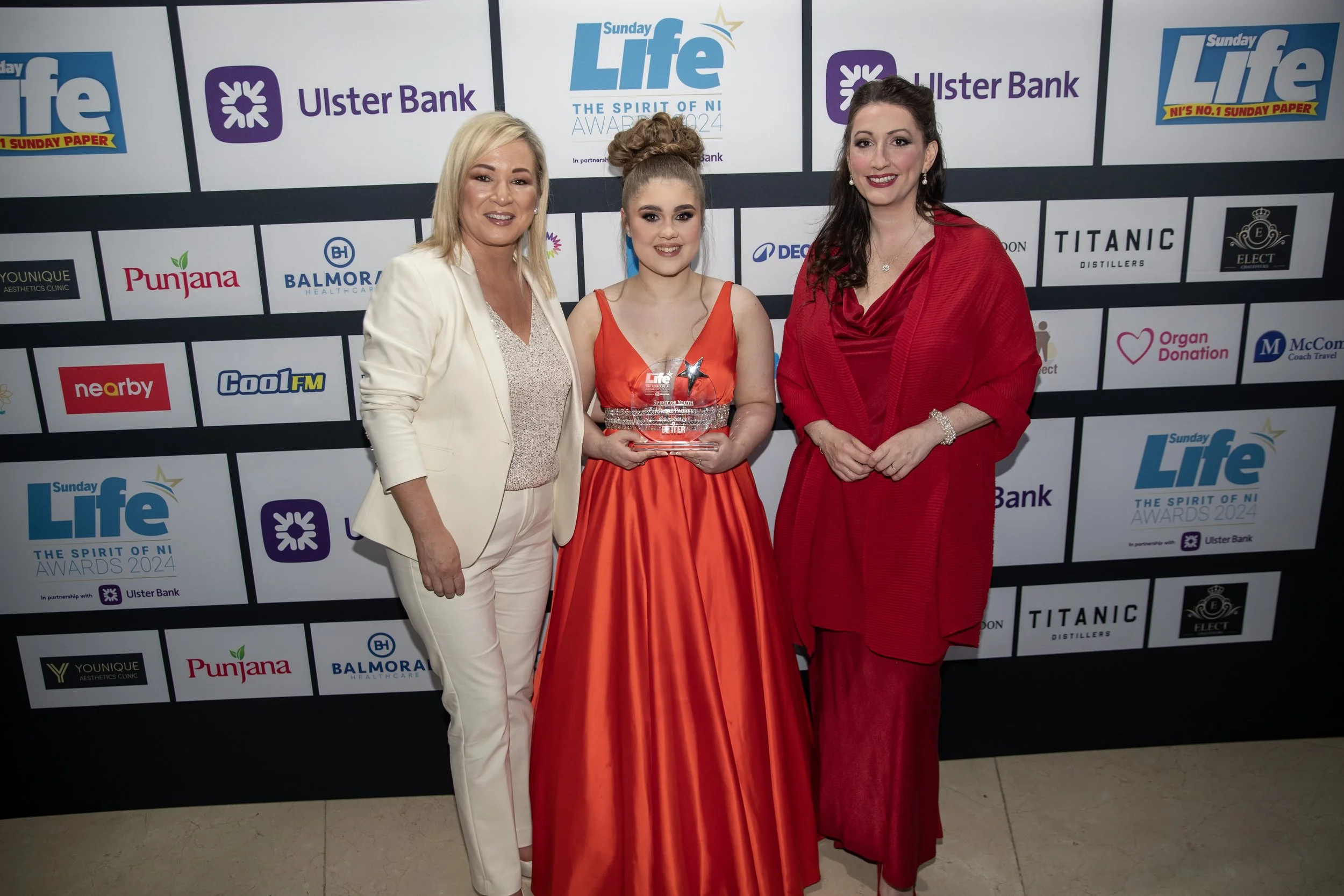 Three women standing together at an awards event. The woman in the middle is holding a trophy and wearing an orange gown. The woman on the left is in a cream-colored suit, and the woman on the right is dressed in a red outfit. They are standing in fr