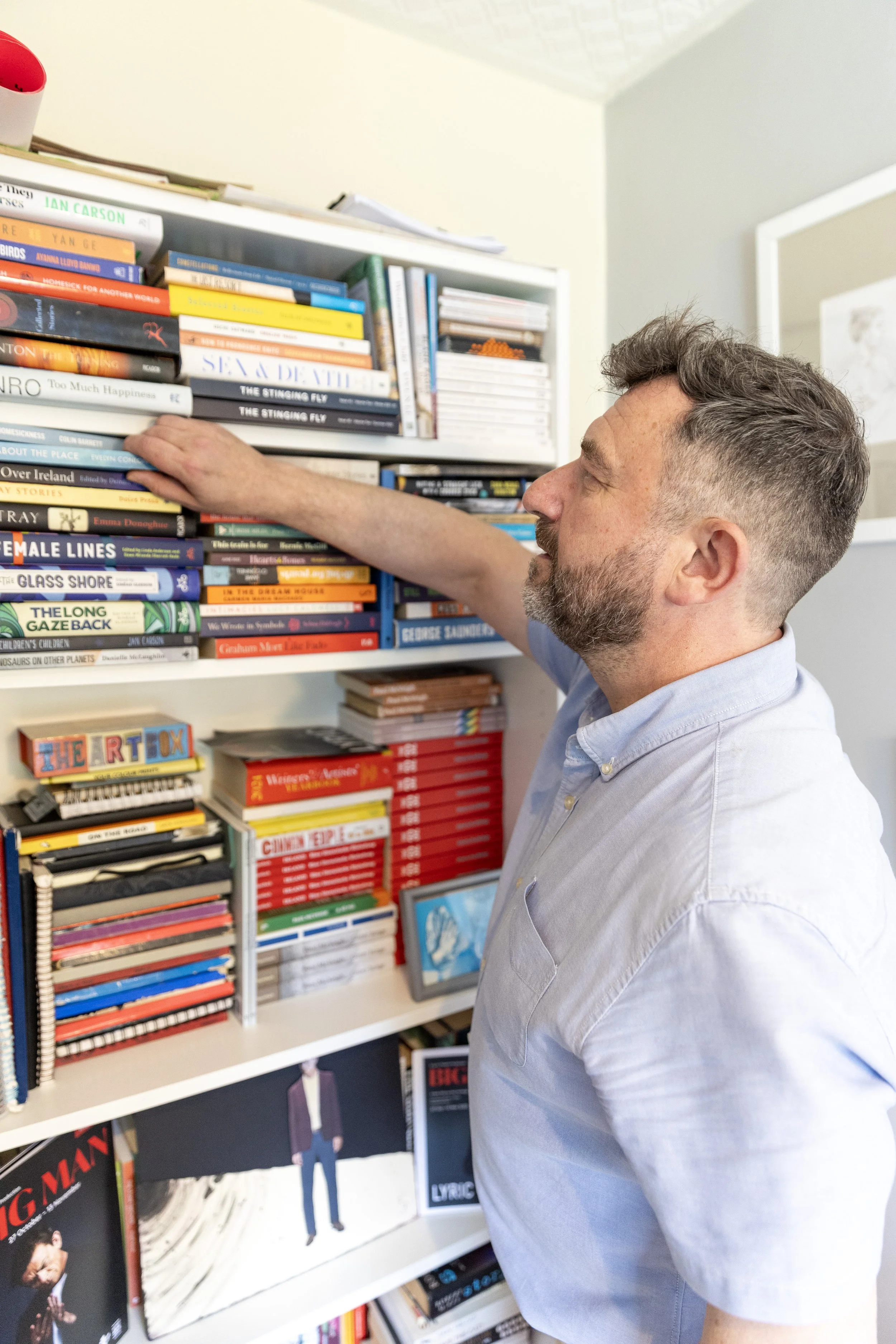 Man selecting a book from a bookshelf filled with various books and magazines in a room.