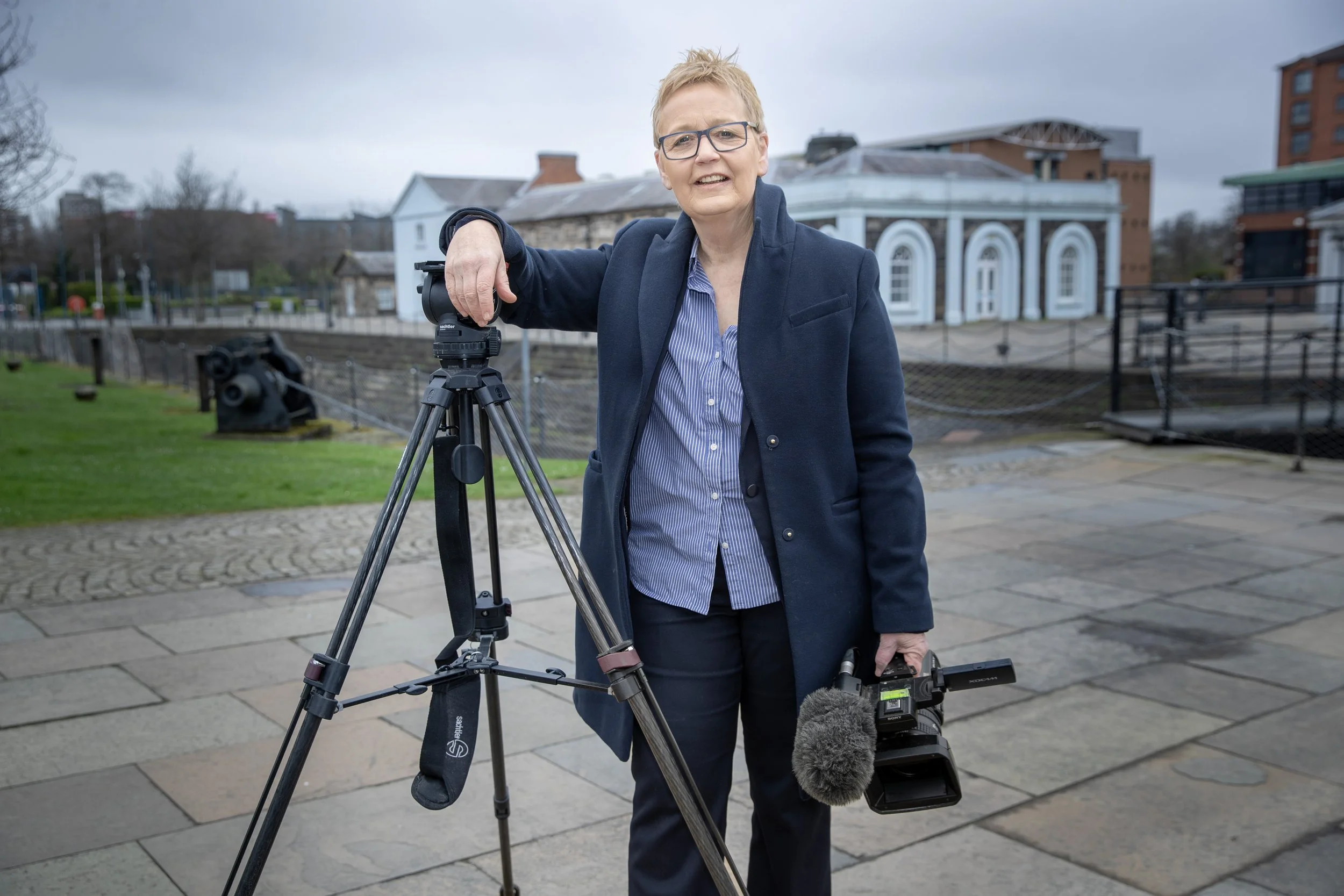 A woman with glasses standing outdoors, holding a professional video camera in her right hand and resting her left arm on a tripod with a camera mounted on it. She is wearing a navy coat and blue shirt. The background shows a historic building, a gra