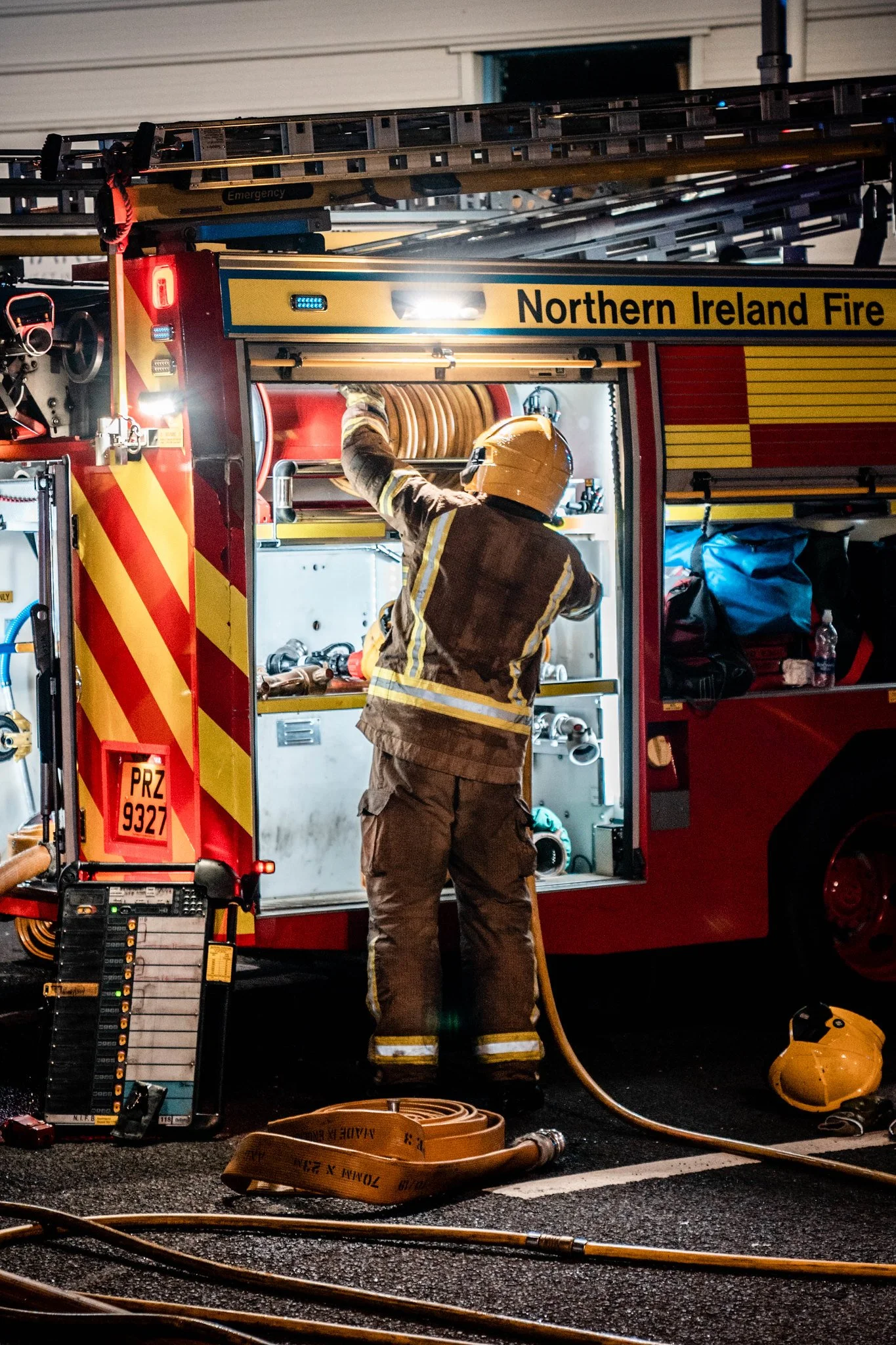 Firefighter in turnout gear organizing equipment inside a Northern Ireland Fire emergency vehicle at night.