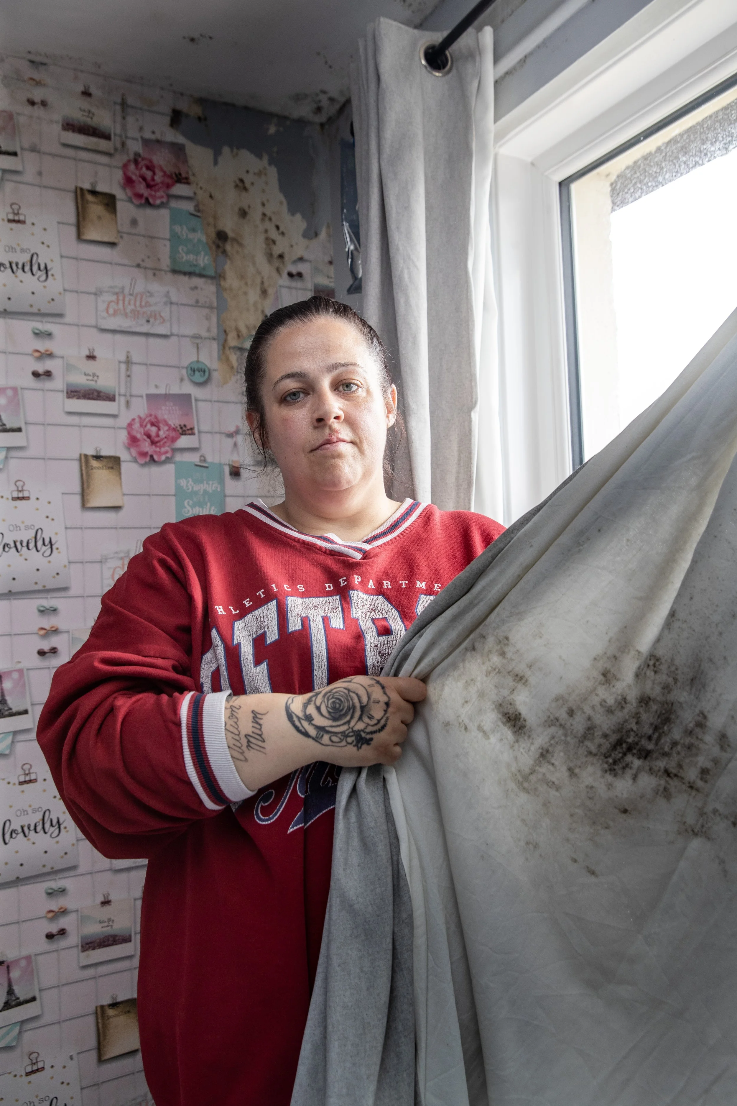 A woman with tattoos on her arm, wearing a red sweatshirt, stands in front of a window with dirty curtains, inside a room with peeling paint and a decorative wall with photographs and notes.