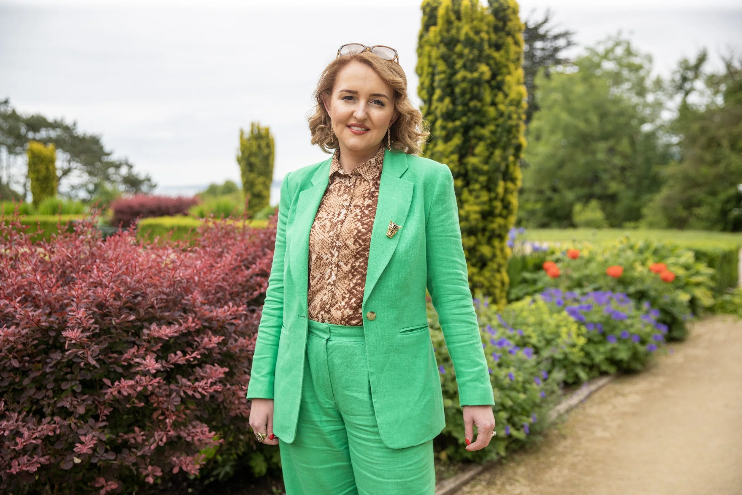 A woman with wavy light brown hair and glasses on her head, wearing a green suit with a brooch, standing in a garden with colorful flowers and greenery.