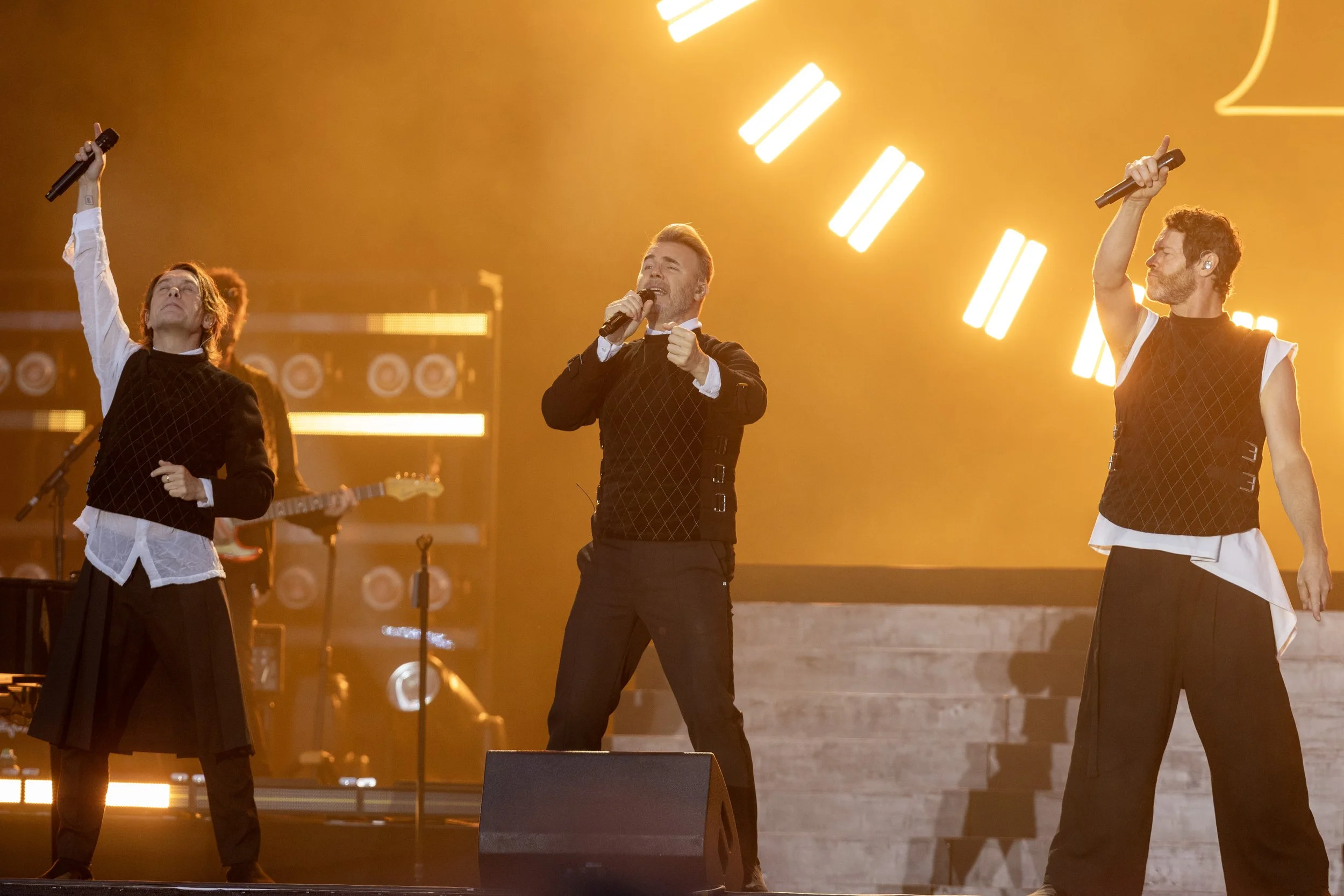 Three male singers performing on stage with bright yellow lights in the background, wearing black and white outfits.