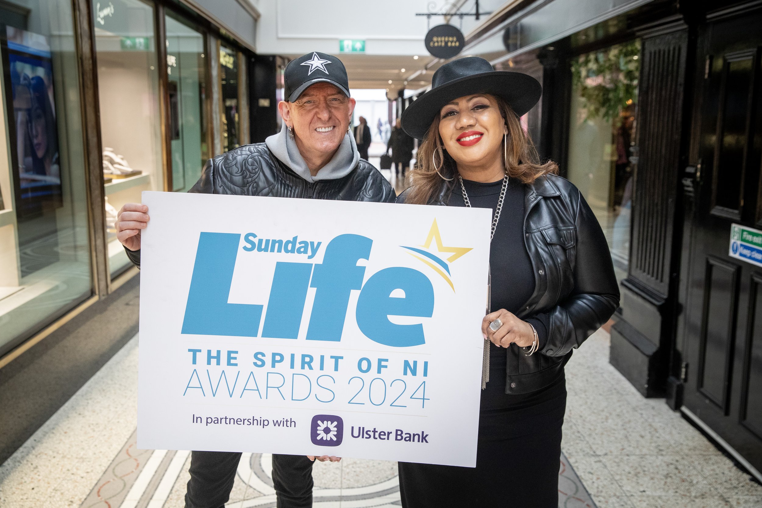 Two people smiling, holding a sign for the Sunday Life Awards 2024 in a shopping mall, with stores and people in the background.