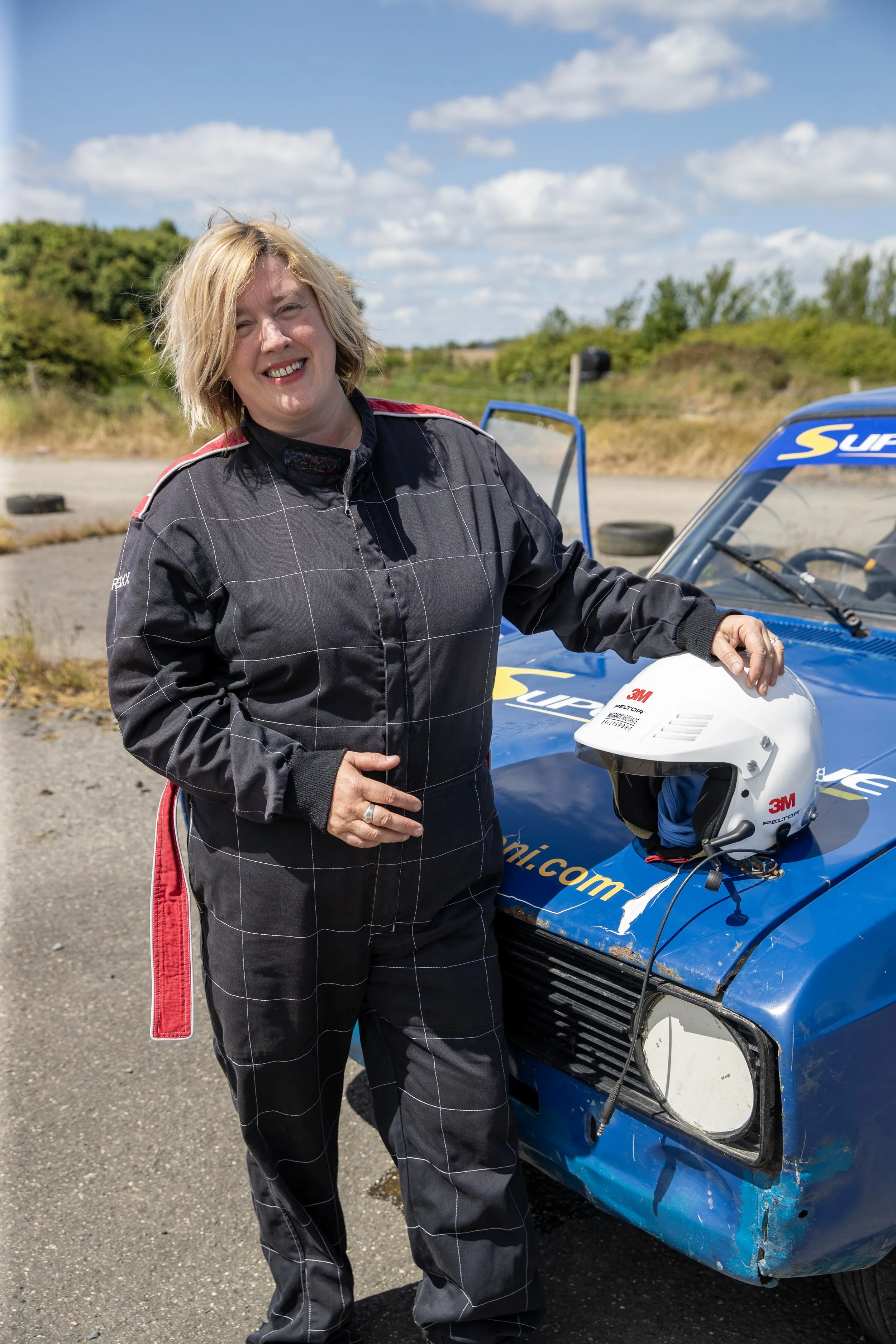 A woman in racing gear standing next to a race car, holding a racing helmet, outdoors under a blue sky with clouds.