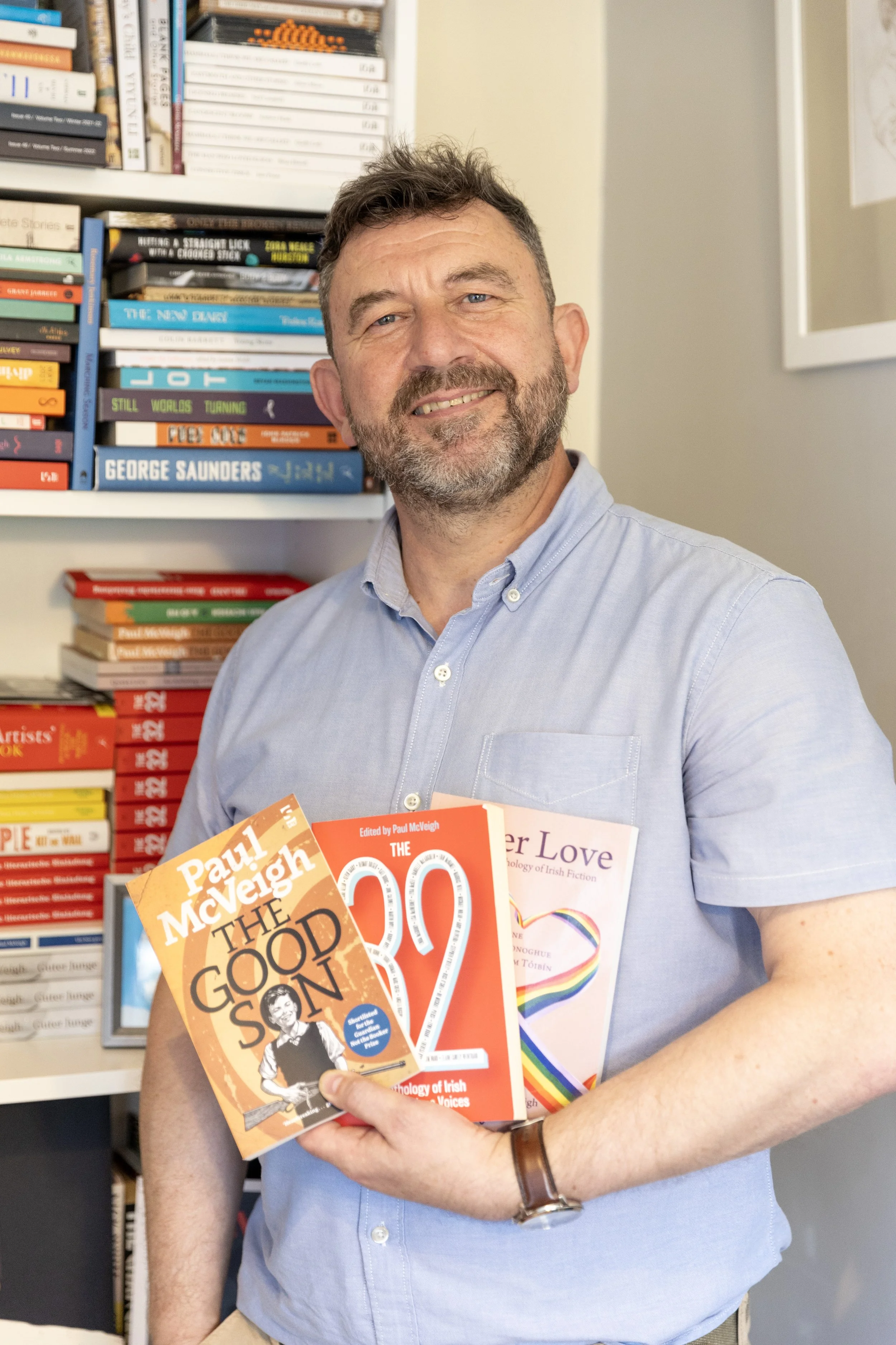 A man with a beard and short hair, wearing a light blue button-up shirt, is standing in front of a bookshelf filled with books. He is smiling and holding three books, one titled 'The Good Son' by Paul McVeigh, one titled 'The 30' edited by Paul McVei