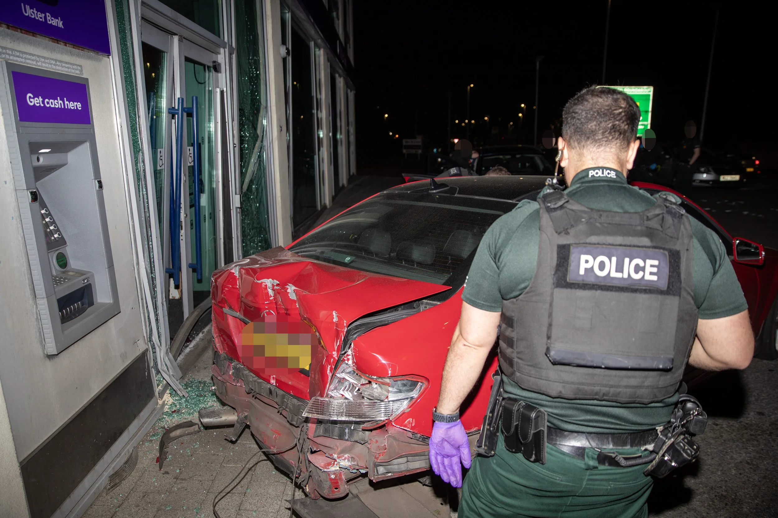 Police officer investigates red car that crashed into an ATM outside a bank at night, causing significant damage to the front of the vehicle.