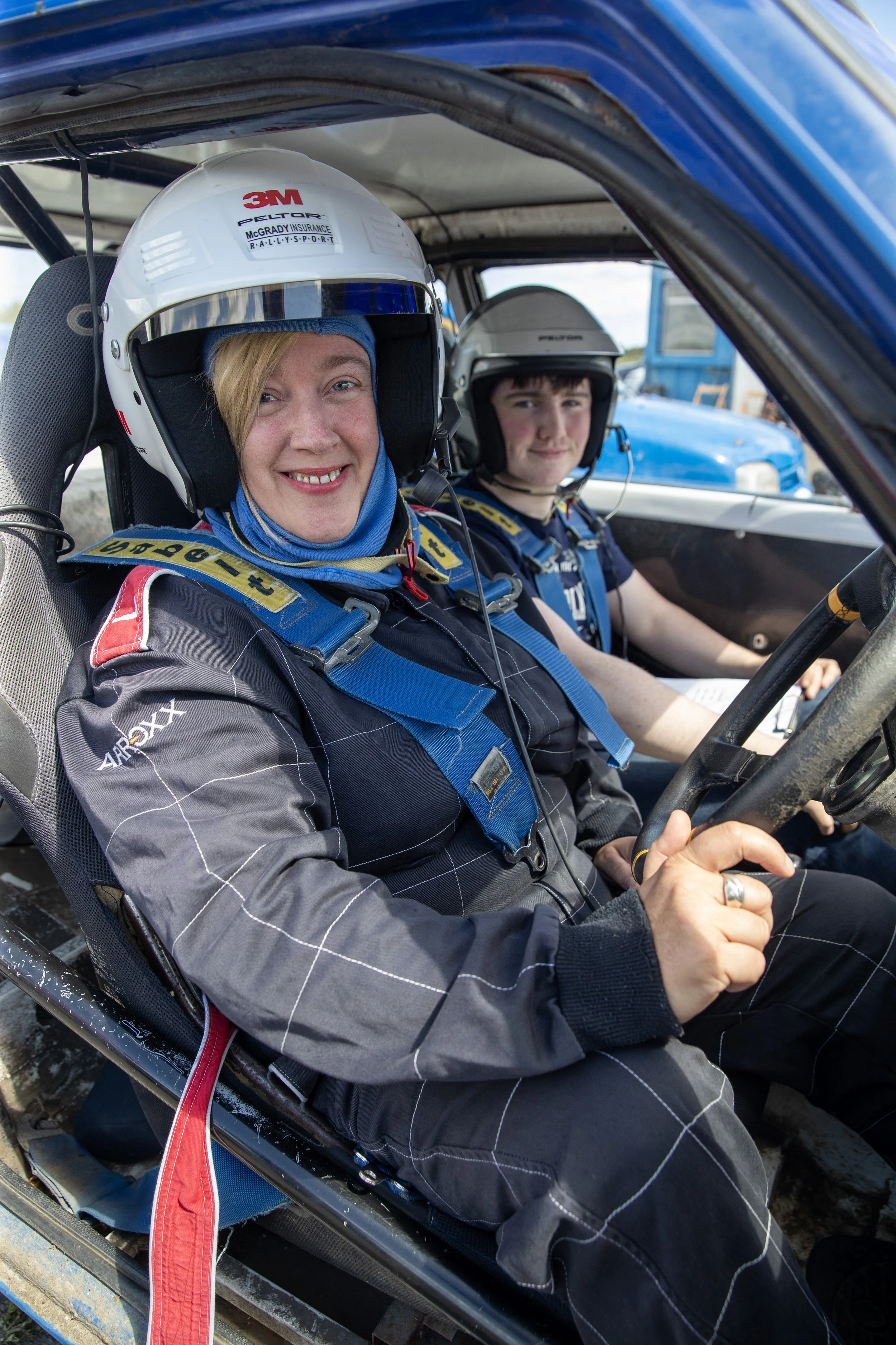 Two women in racing helmets and suits sitting in a race car, smiling, with a male passenger in the background.