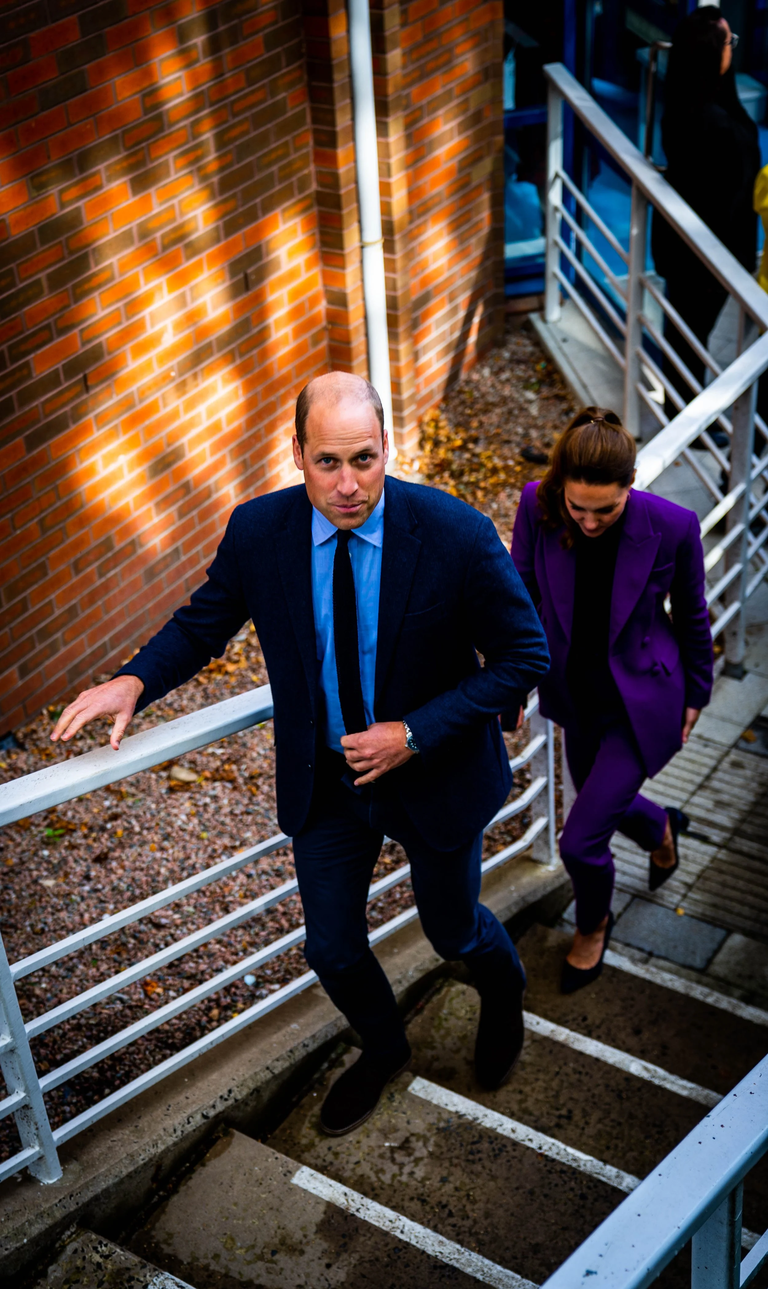 A man in a suit and a woman in a purple suit walking up outdoor stairs beside a brick wall.