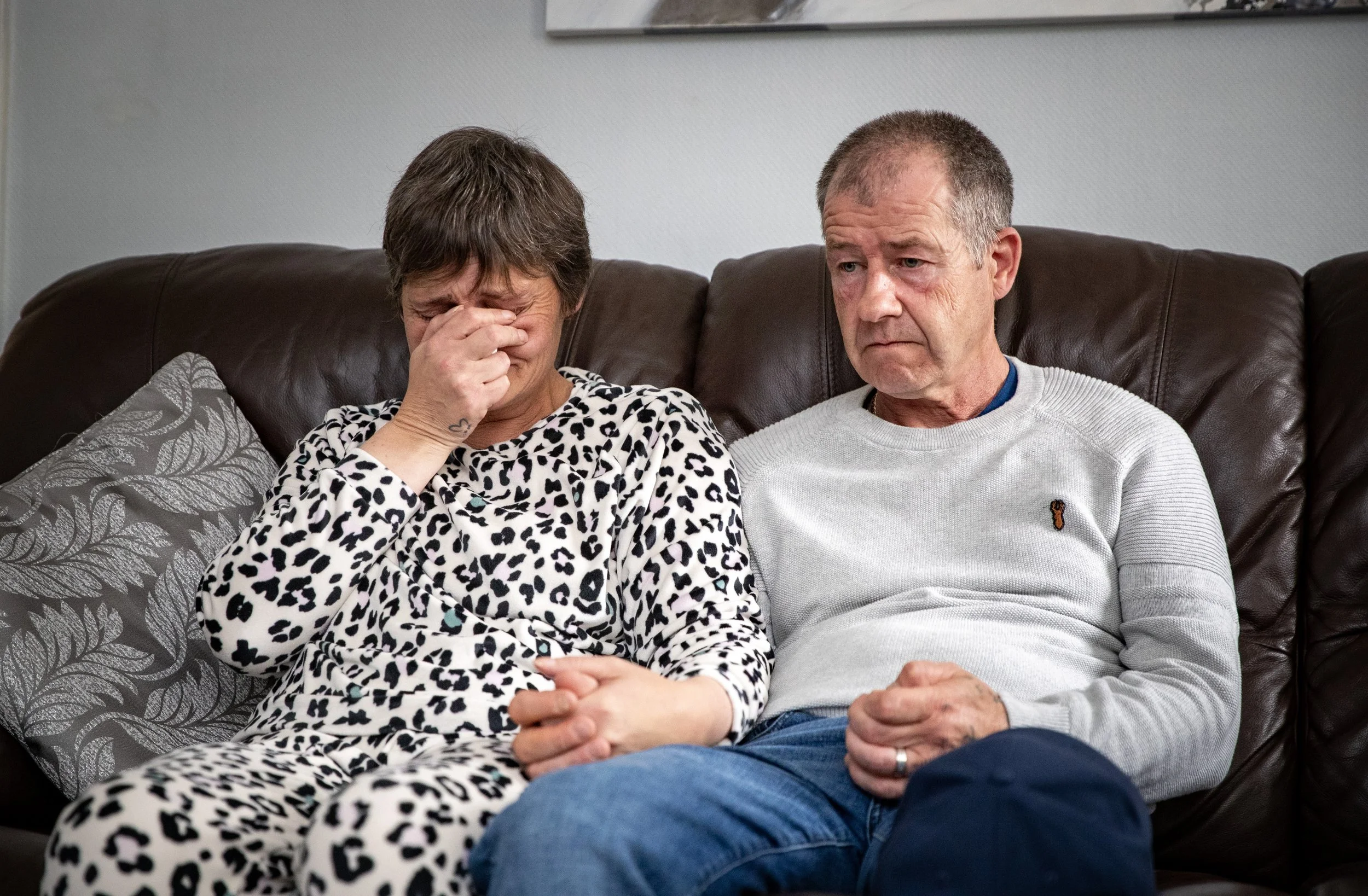 A woman with short dark hair and a woman with short gray hair sitting on a brown leather couch, visibly distressed, with the woman on the left holding her face and the man on the right looking serious.