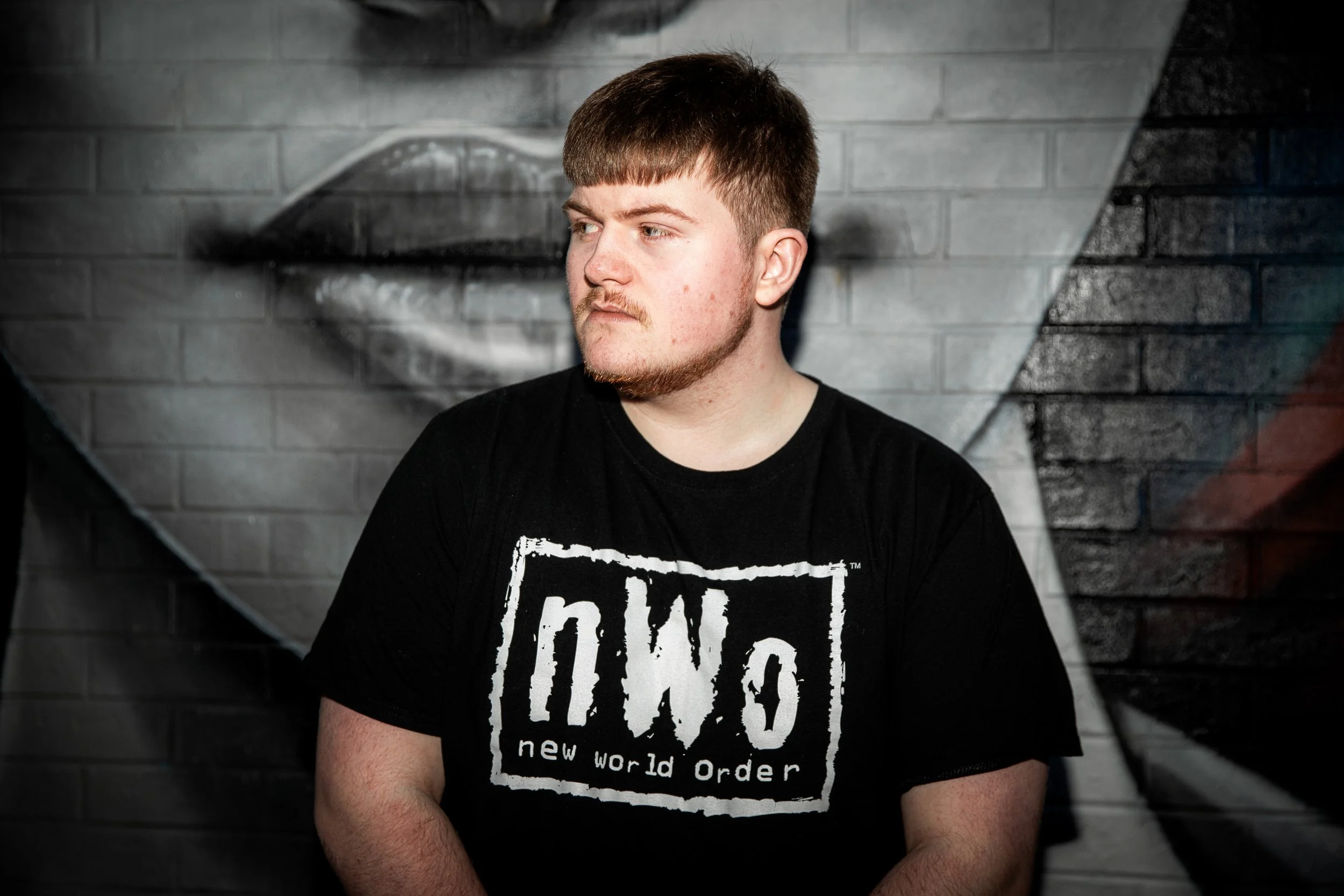 A young man with short brown hair and a beard, wearing a black t-shirt with the text "nWo new world order," is standing in front of a graffiti-style mural of lips on a brick wall.