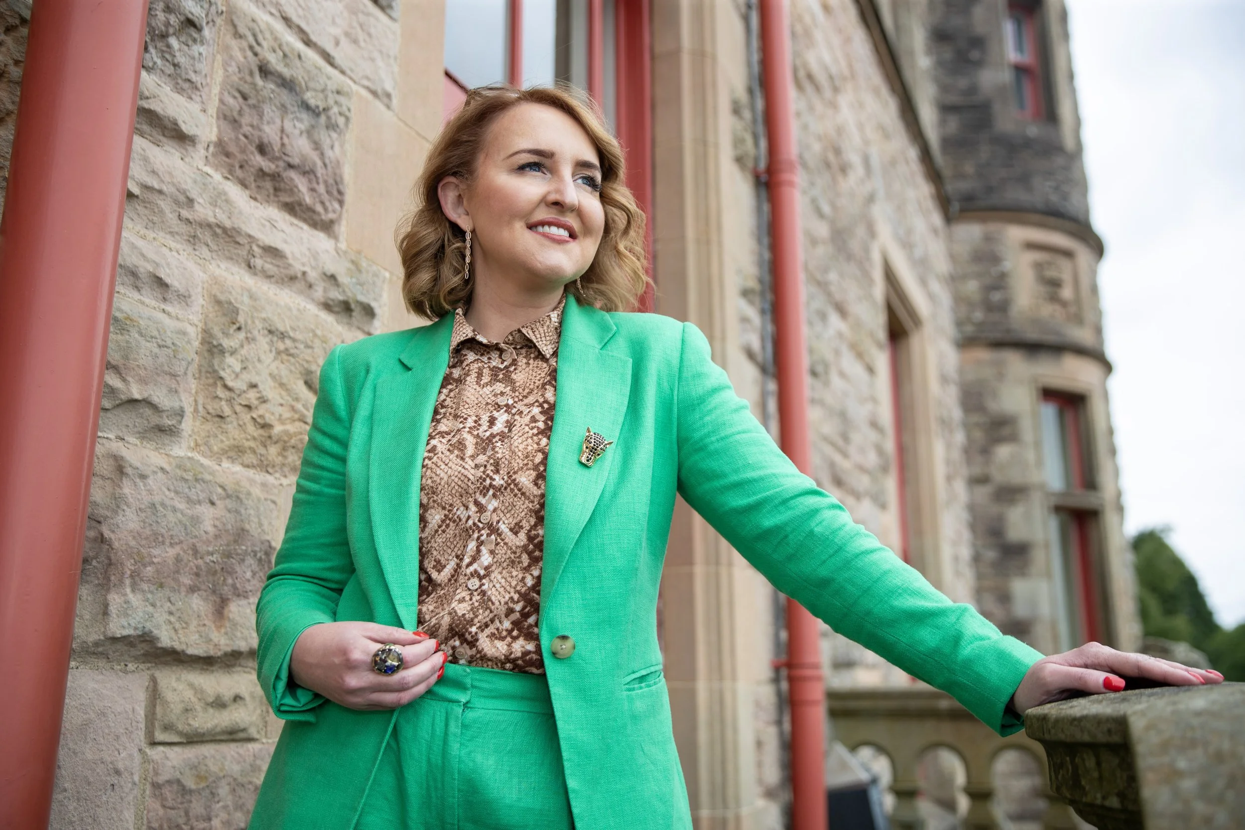 A woman with shoulder-length wavy hair, wearing a bright green suit and a patterned blouse, standing outside a stone building with her hand on a stone ledge and smiling.