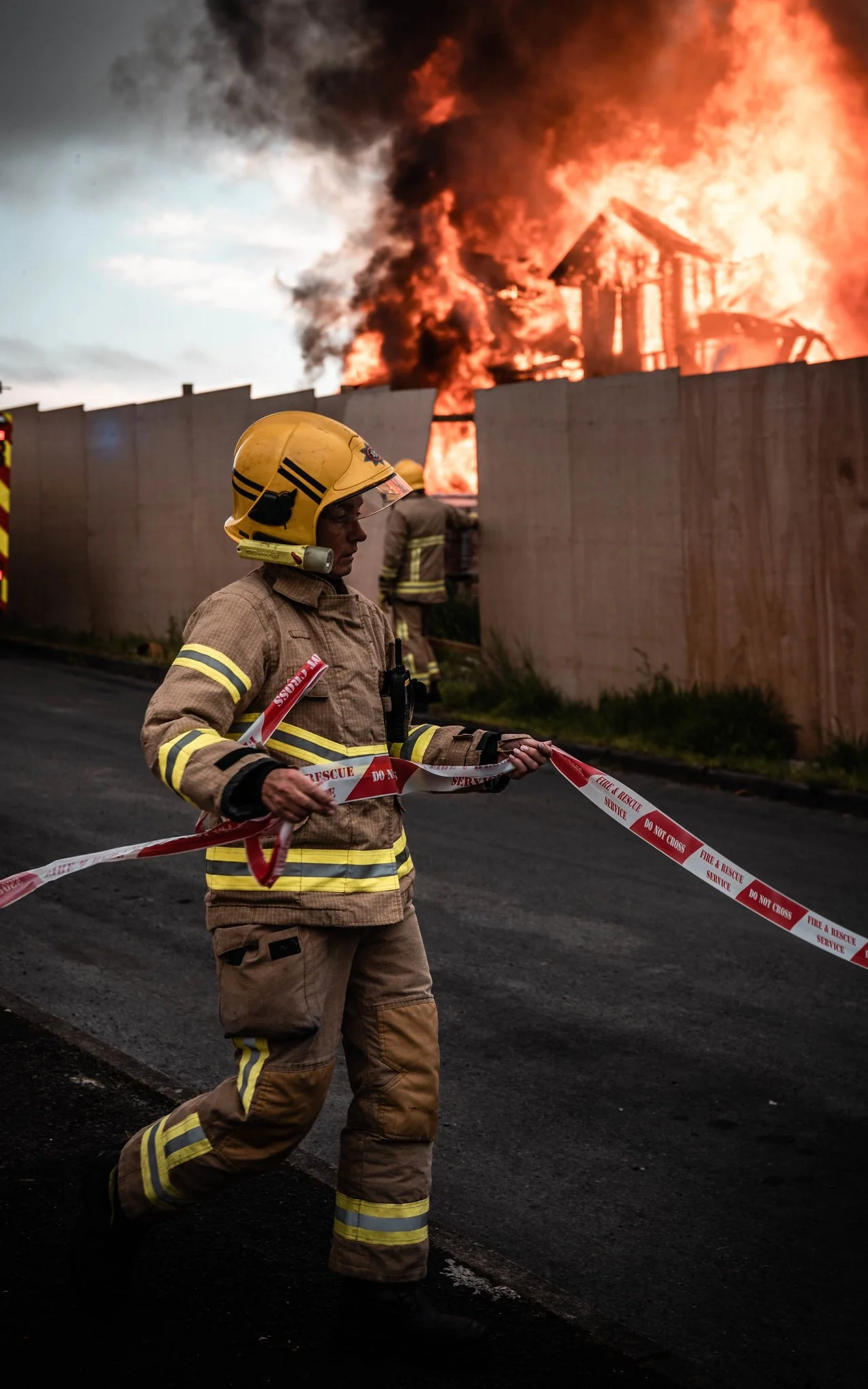 A firefighter in beige turnout gear and helmet holding fire rescue tape, standing on a street as a house burns intensely behind a barrier wall, with black smoke and flames visible.