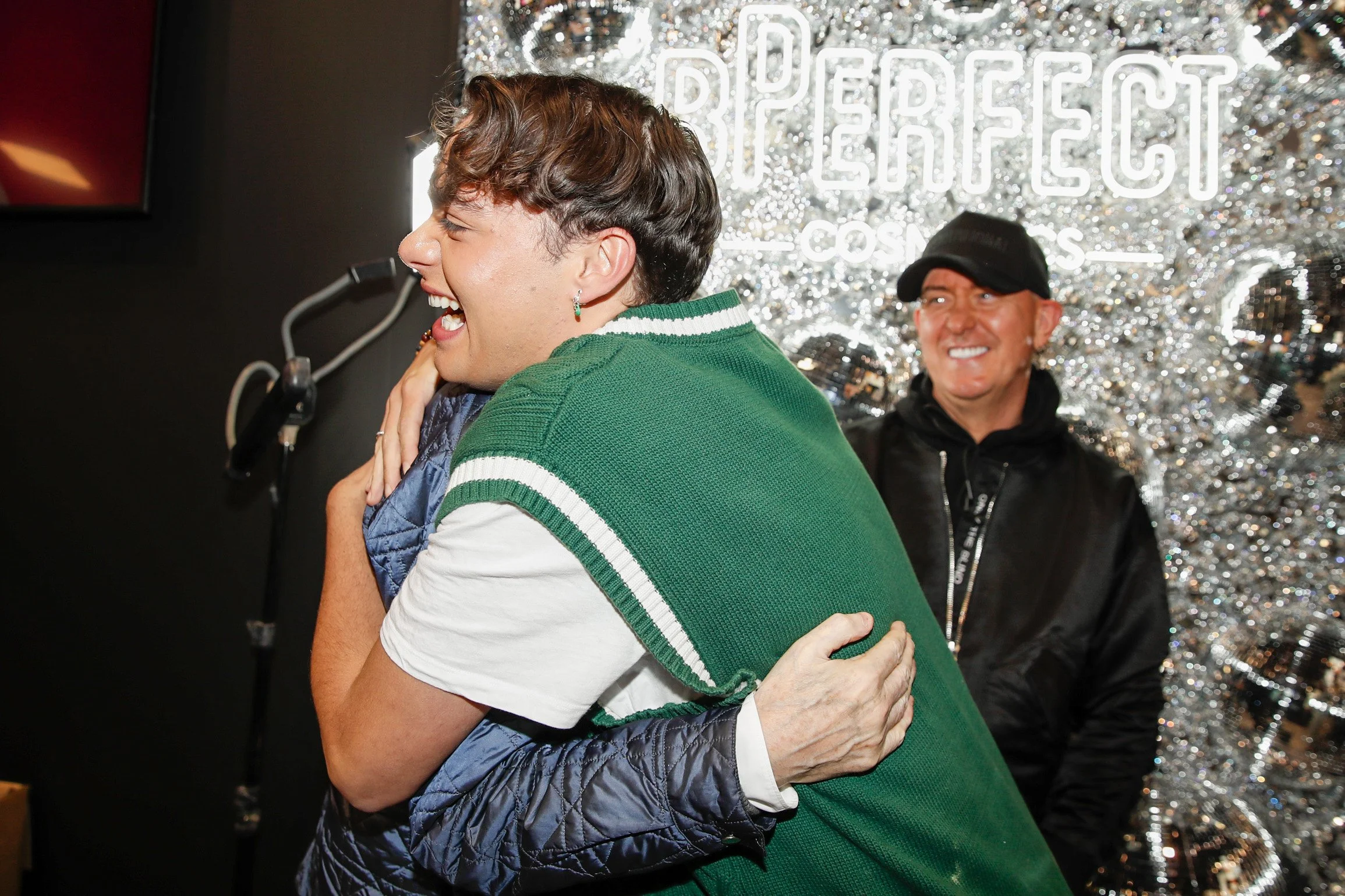 Two people hugging and smiling, with a cheerful woman on the left and a man in the background, in front of a shiny, decorative backdrop that says "Perfect".
