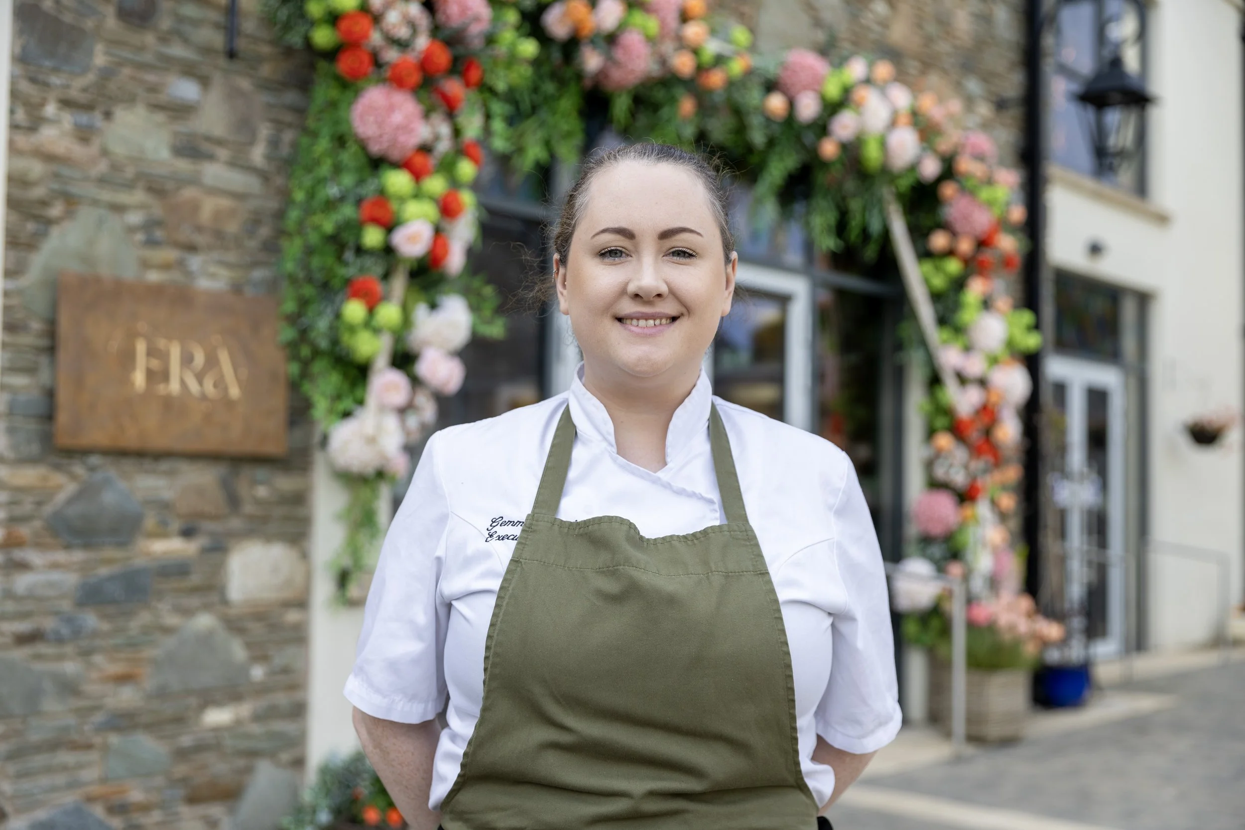 A smiling woman in a white chef's coat and green apron standing in front of a flower-decorated building.
