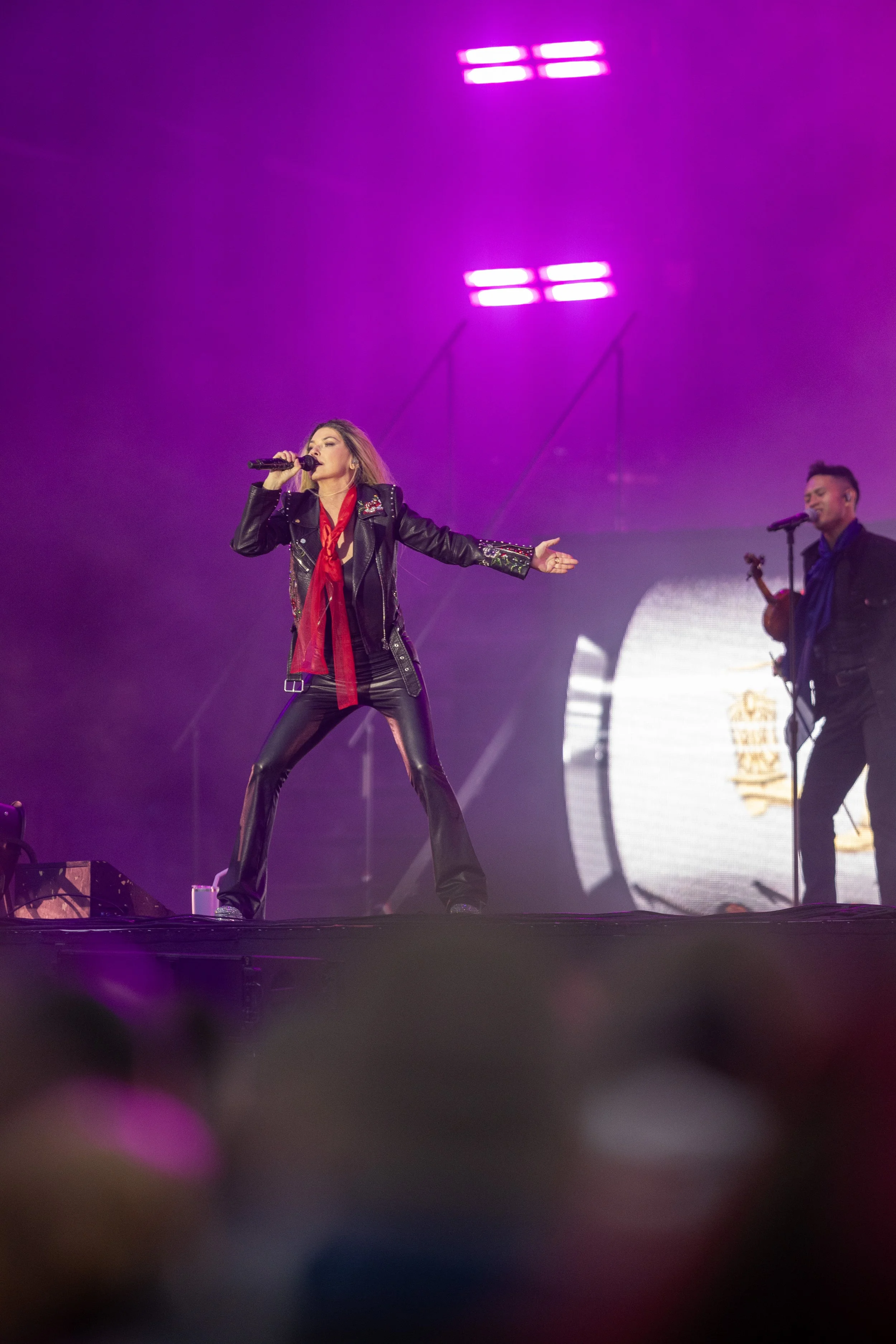 A female singer performs on stage with purple lighting in the background, wearing a leather jacket, black leather pants, and a red scarf, holding a microphone in her right hand.
