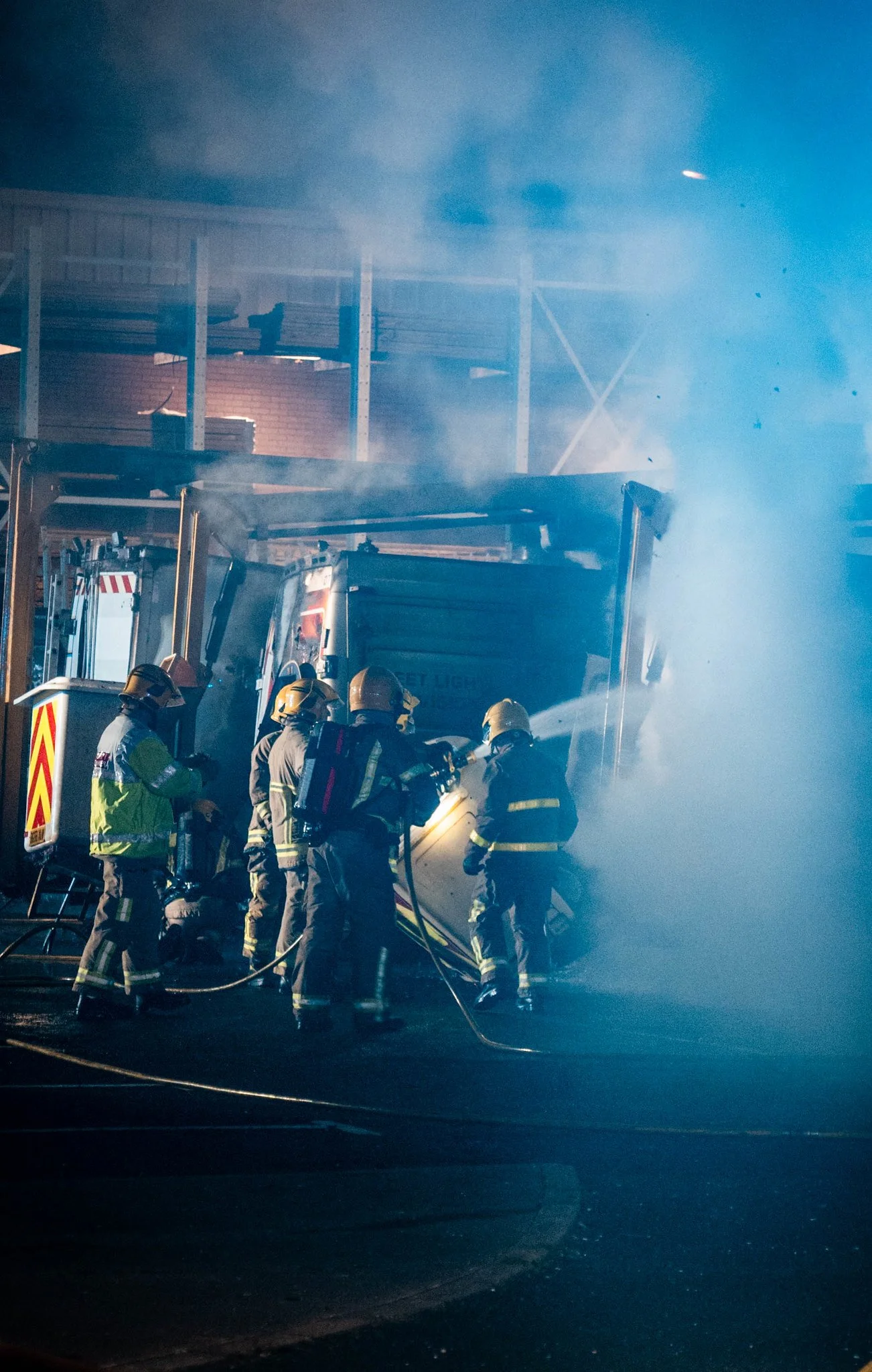 Firefighters battling a warehouse fire at night, with smoke and water spray visible.