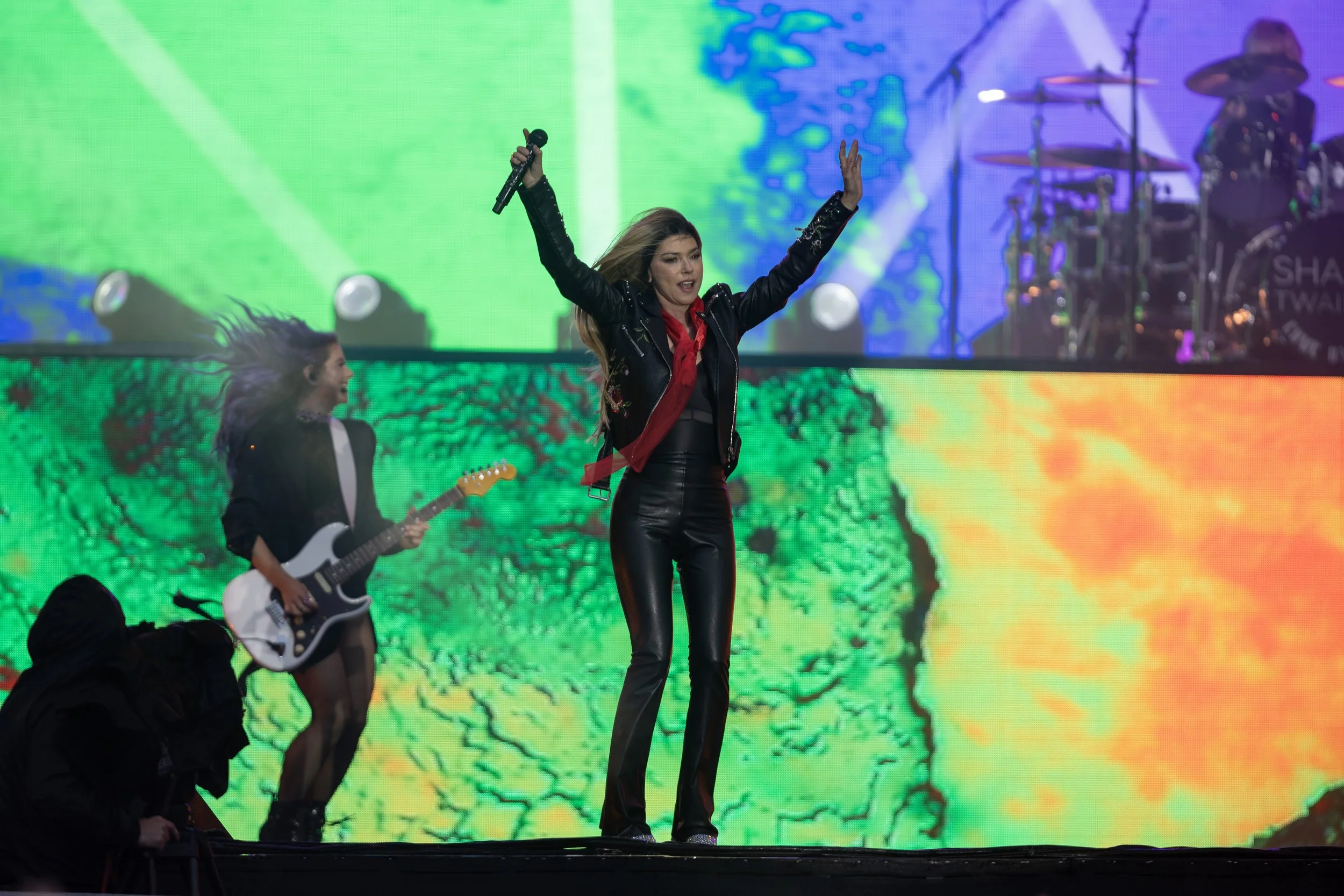 Female performer singing on stage with her arms raised, wearing black leather pants and a black jacket, with a colorful digital background and a drummer in the background.