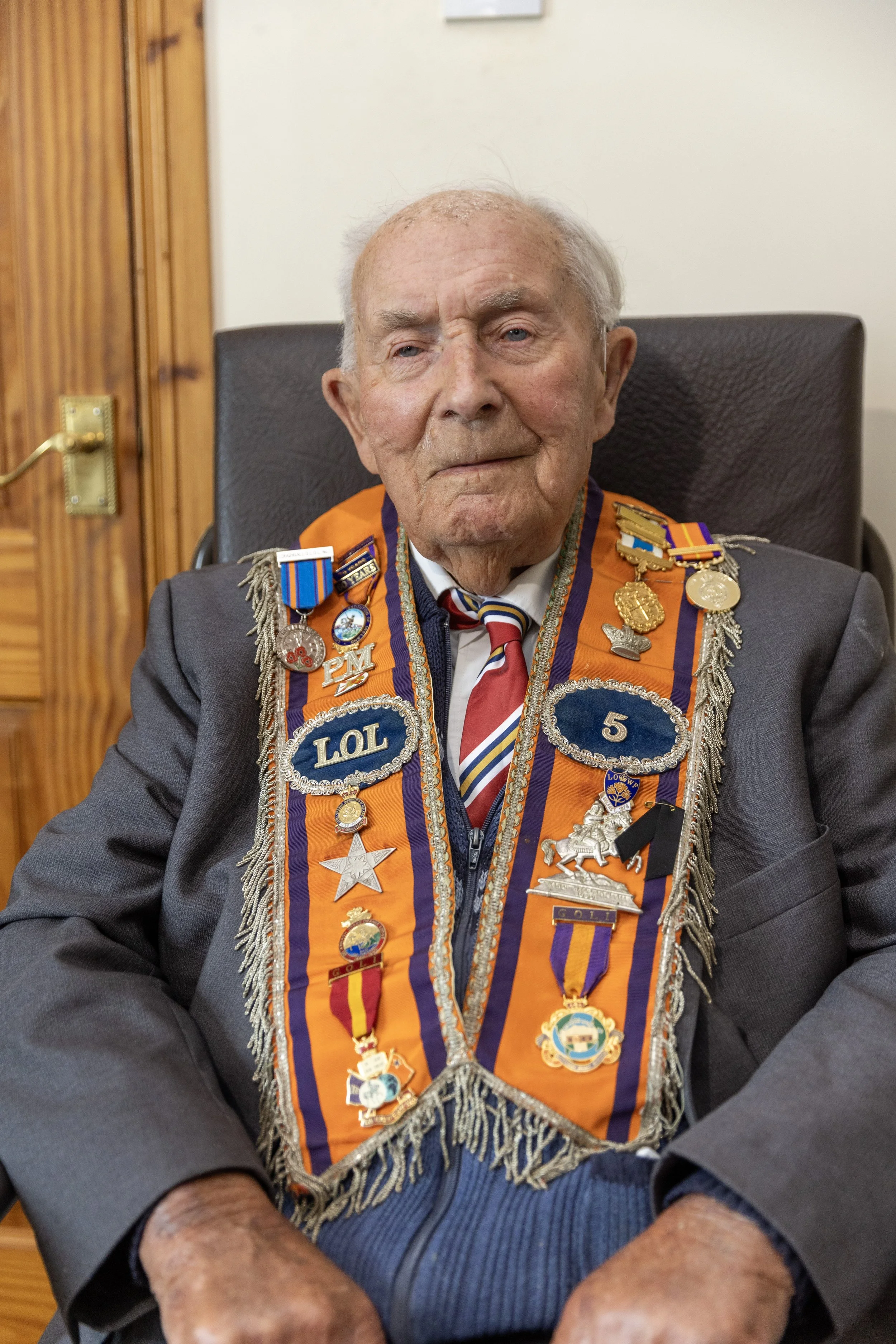 An elderly man wearing a suit and a decorated sash with various medals and pins standing for a portrait.