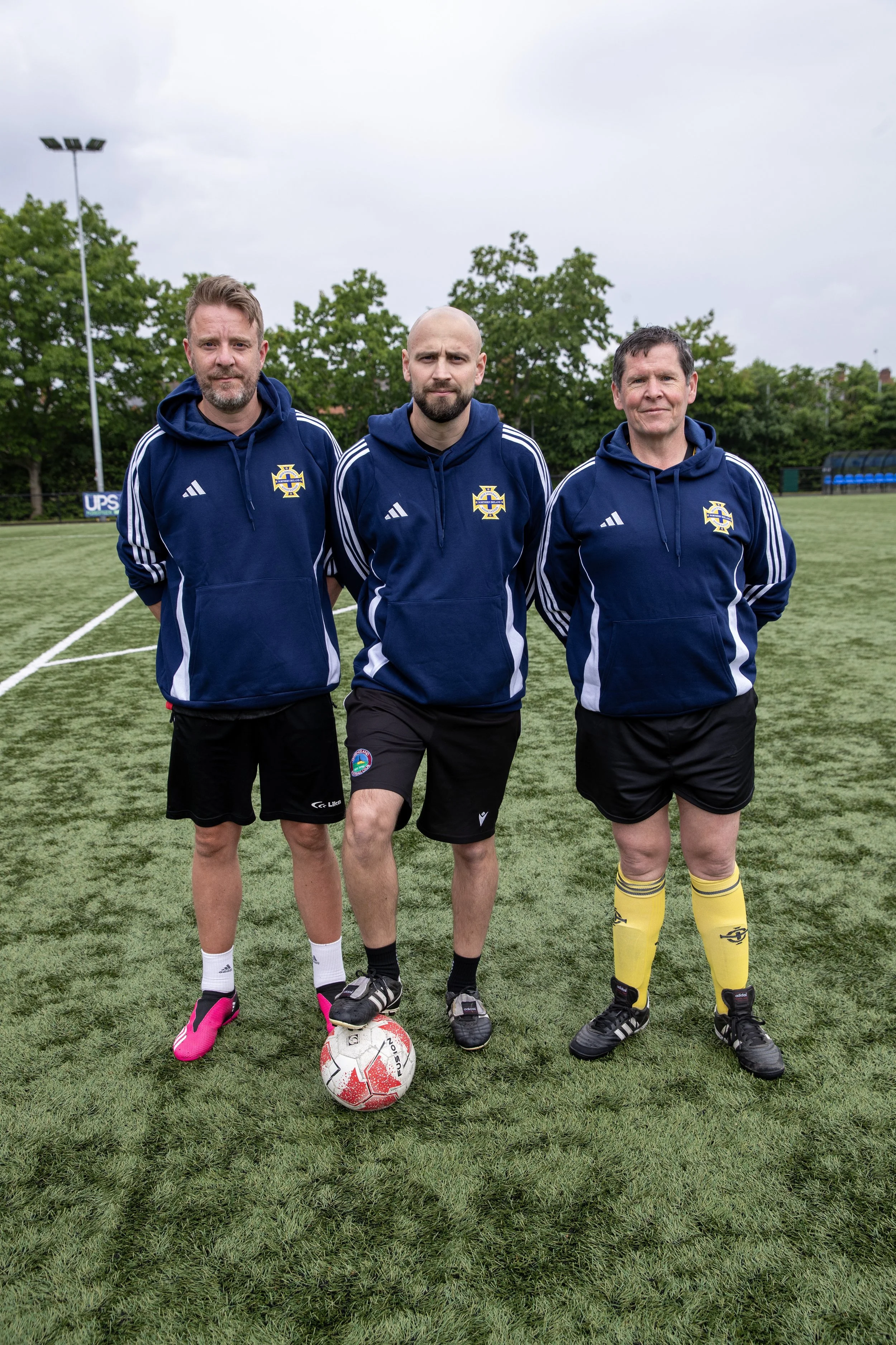 Three men standing on a soccer field wearing matching blue hoodies and shorts, with a soccer ball at their feet, posing for a photo
