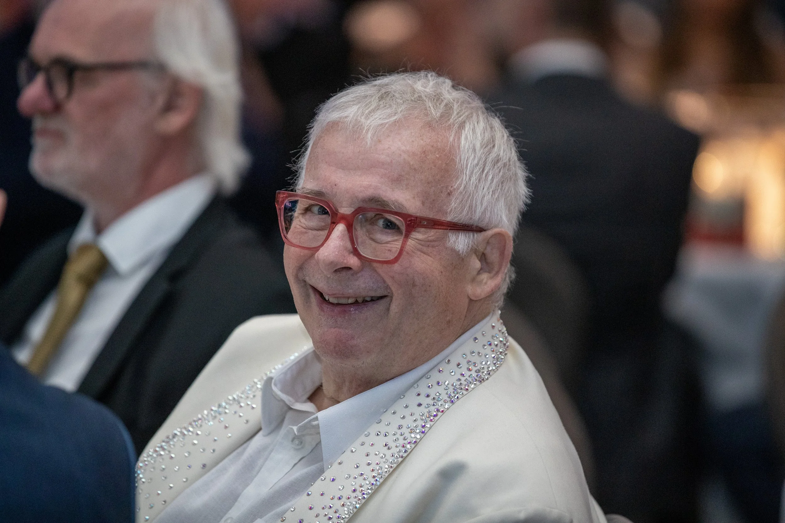 A smiling elderly man with white hair and red glasses, wearing a white blazer with rhinestones on the lapels, sitting at an event surrounded by other guests.