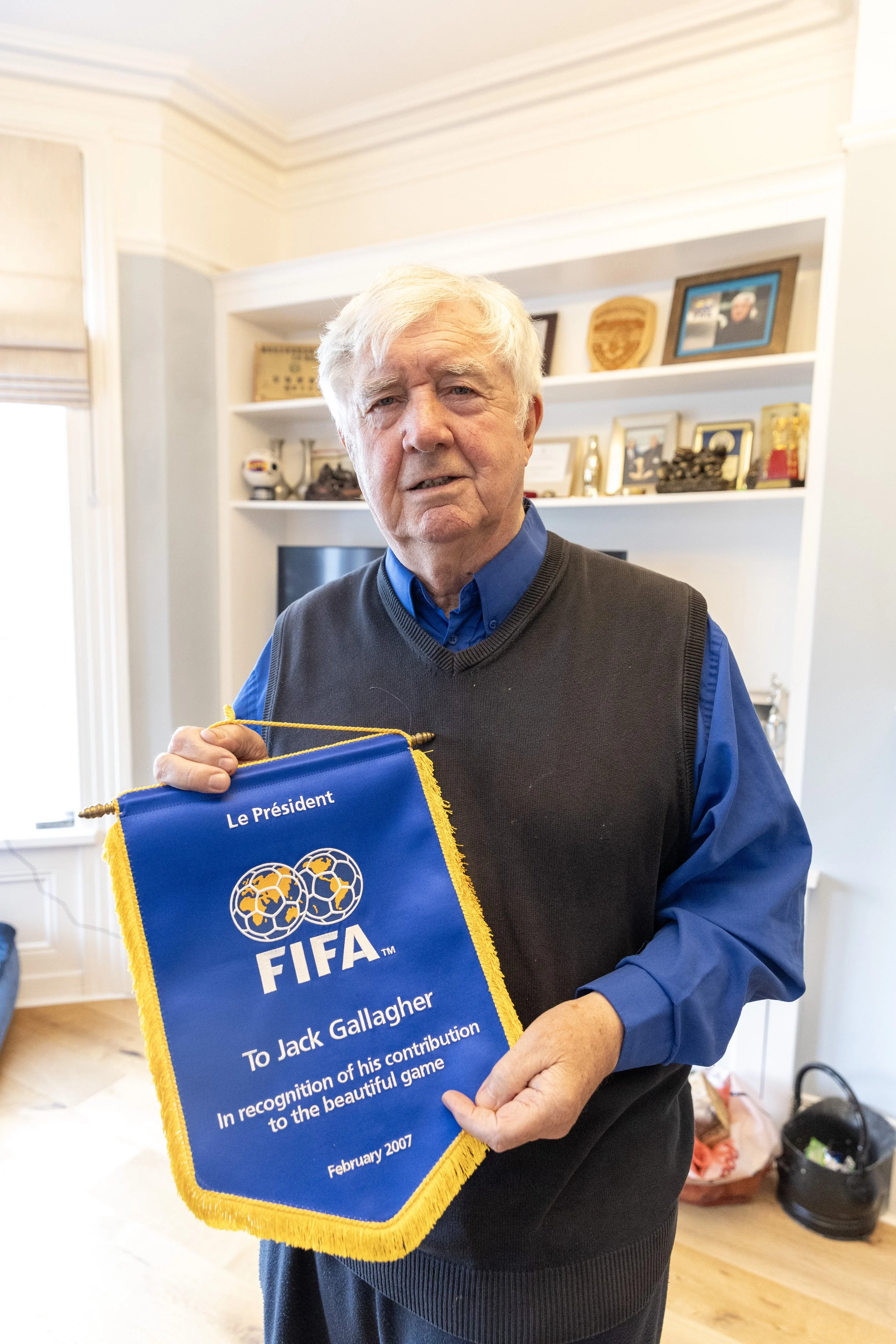 An elderly man holding a blue FIFA plaque with gold fringe, recognizing Jack Gallagher's contribution to soccer in February 2007, inside a room with framed photos and awards on the shelves behind him.