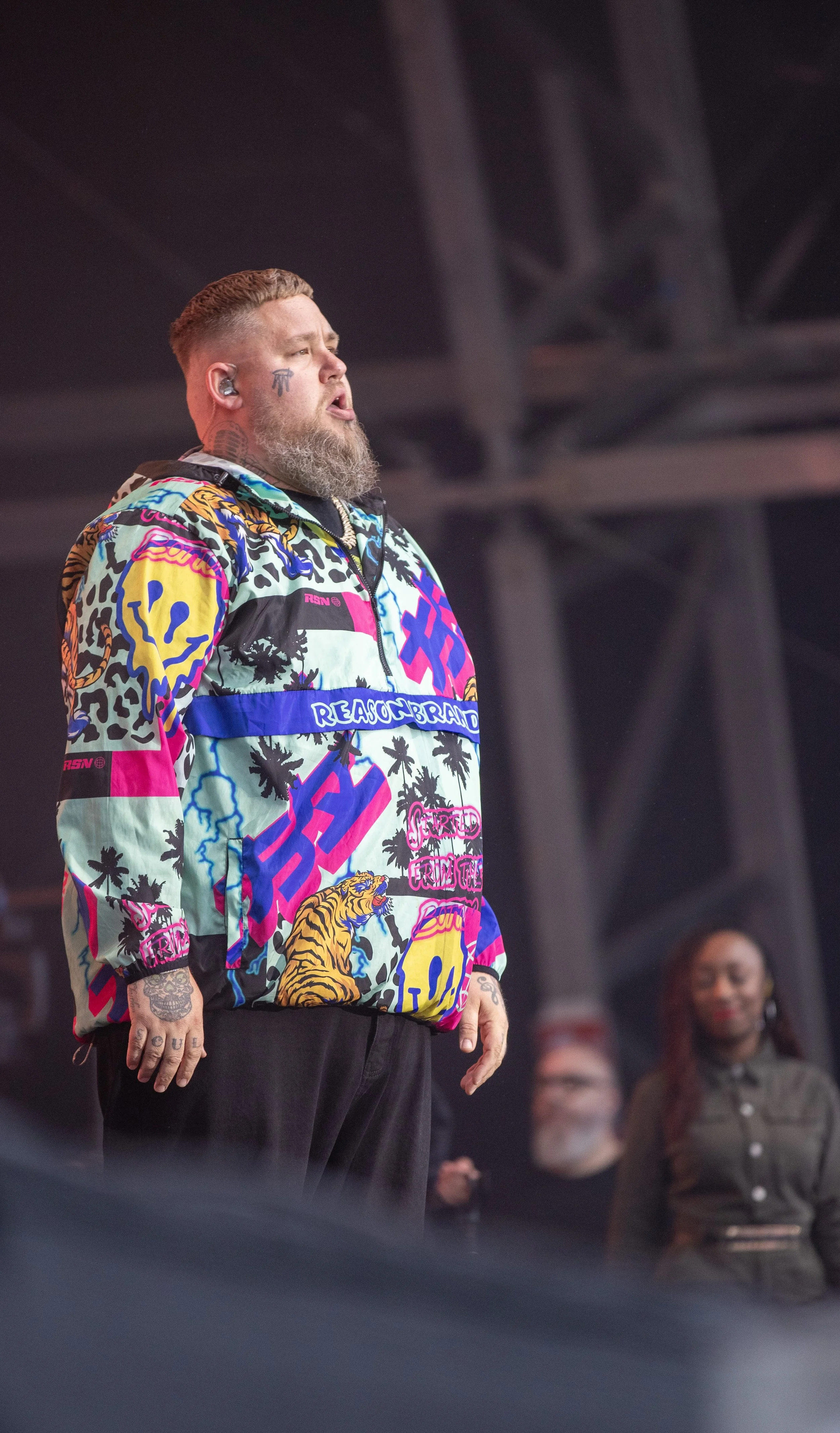 Man with tattoos and a beard wearing a colorful jacket with tiger, skull, and leopard print patterns, standing on stage with a serious expression.