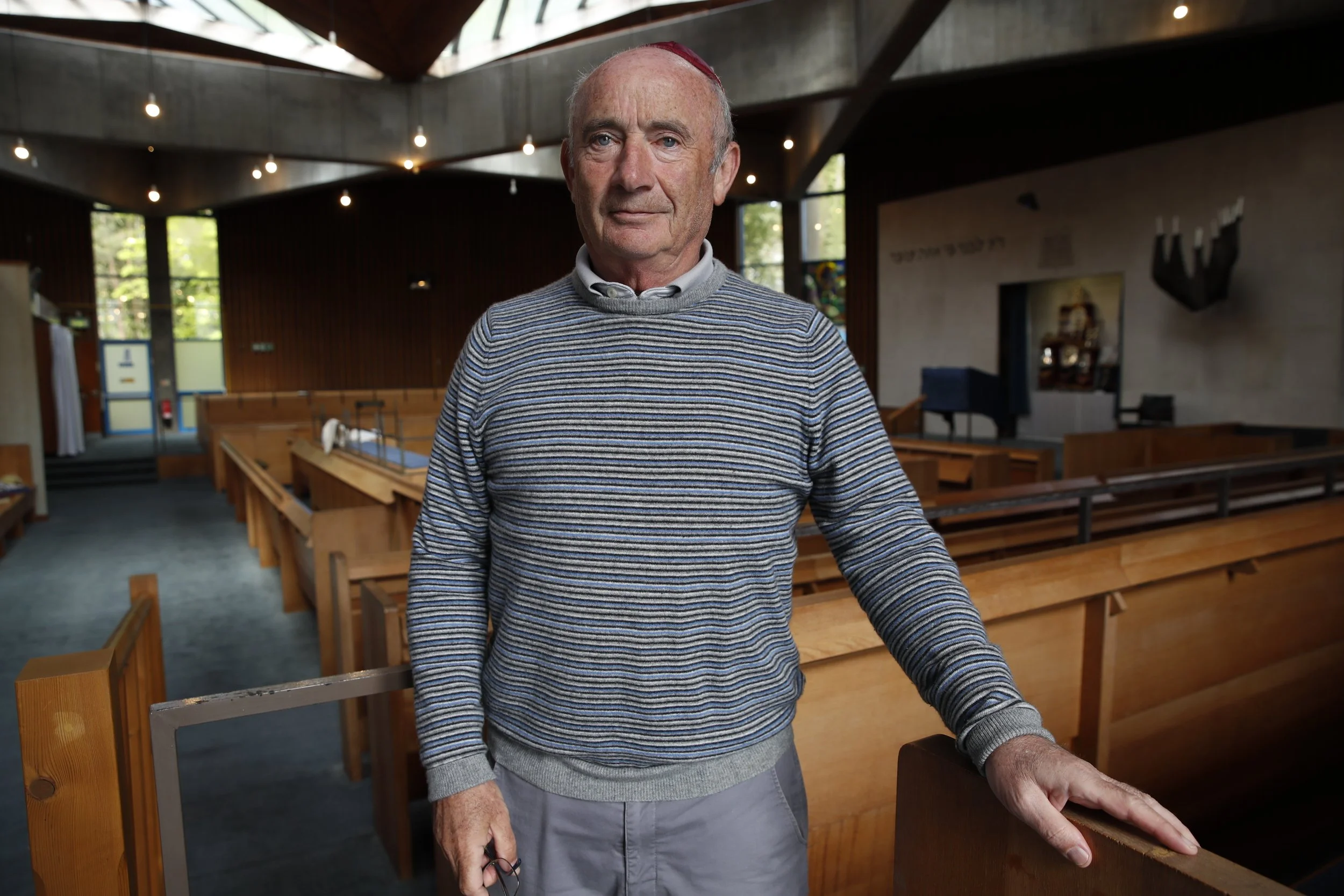 An elderly man standing inside a modern church with wooden pews, wearing a striped sweater and holding glasses, looking at the camera.
