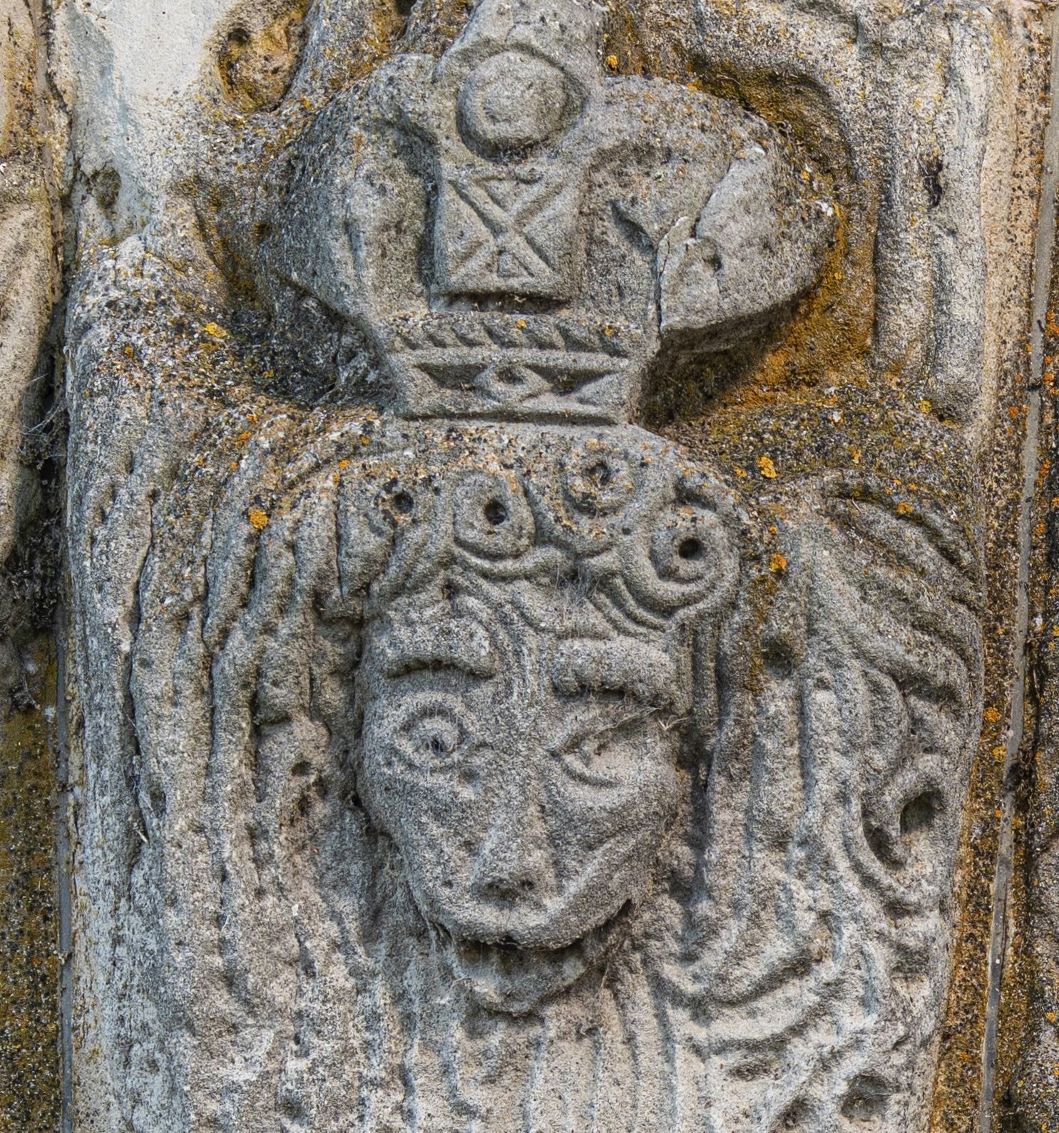 Close-up of an ancient stone carving featuring a humanoid face with exaggerated features, surrounded by intricate patterns and symbols, with moss and lichen growing on the surface.