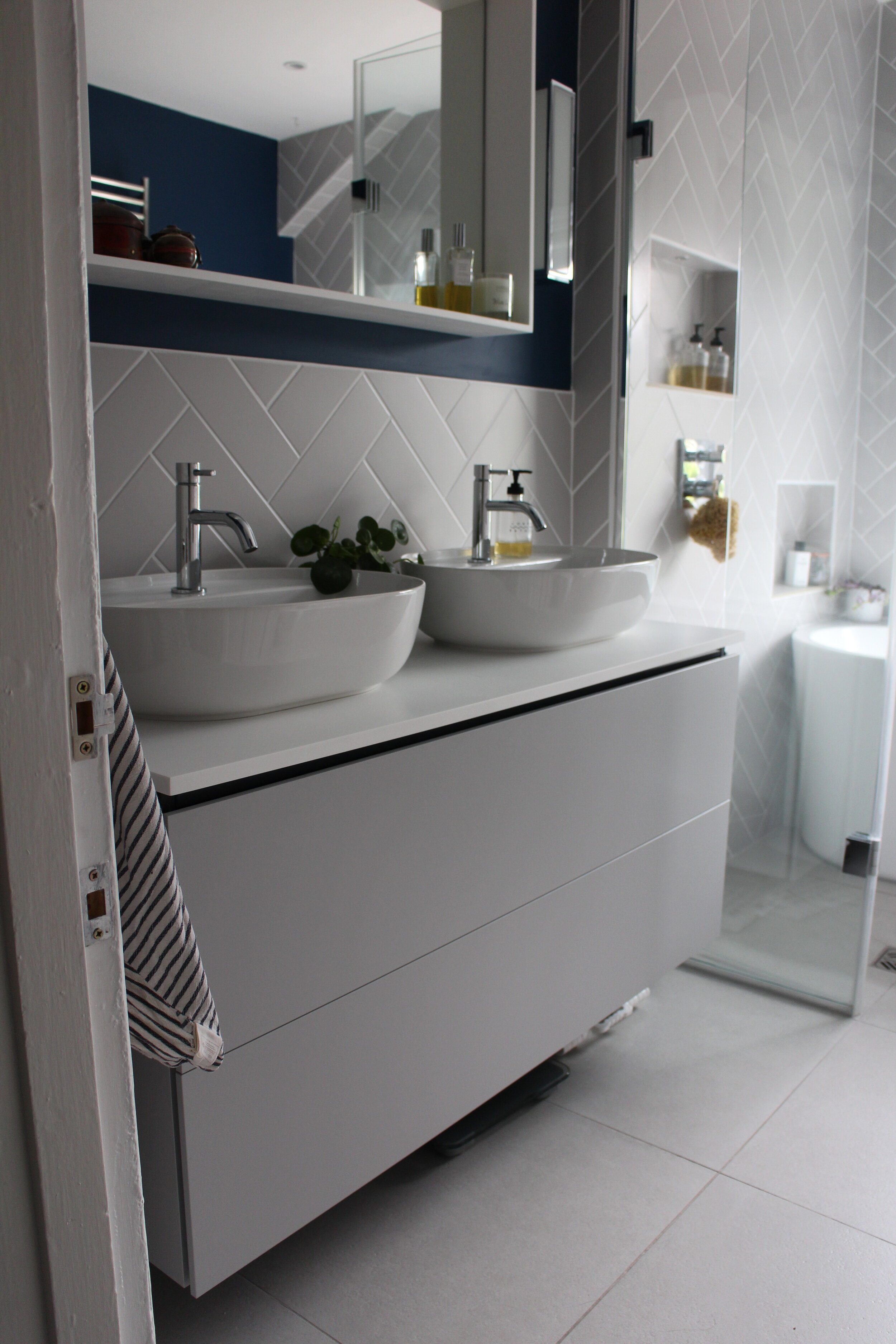 Modern bathroom with a double sink vanity, large mirror, and glass shower enclosure. Decor includes soap dispensers, a plant, and bottles of toiletries.