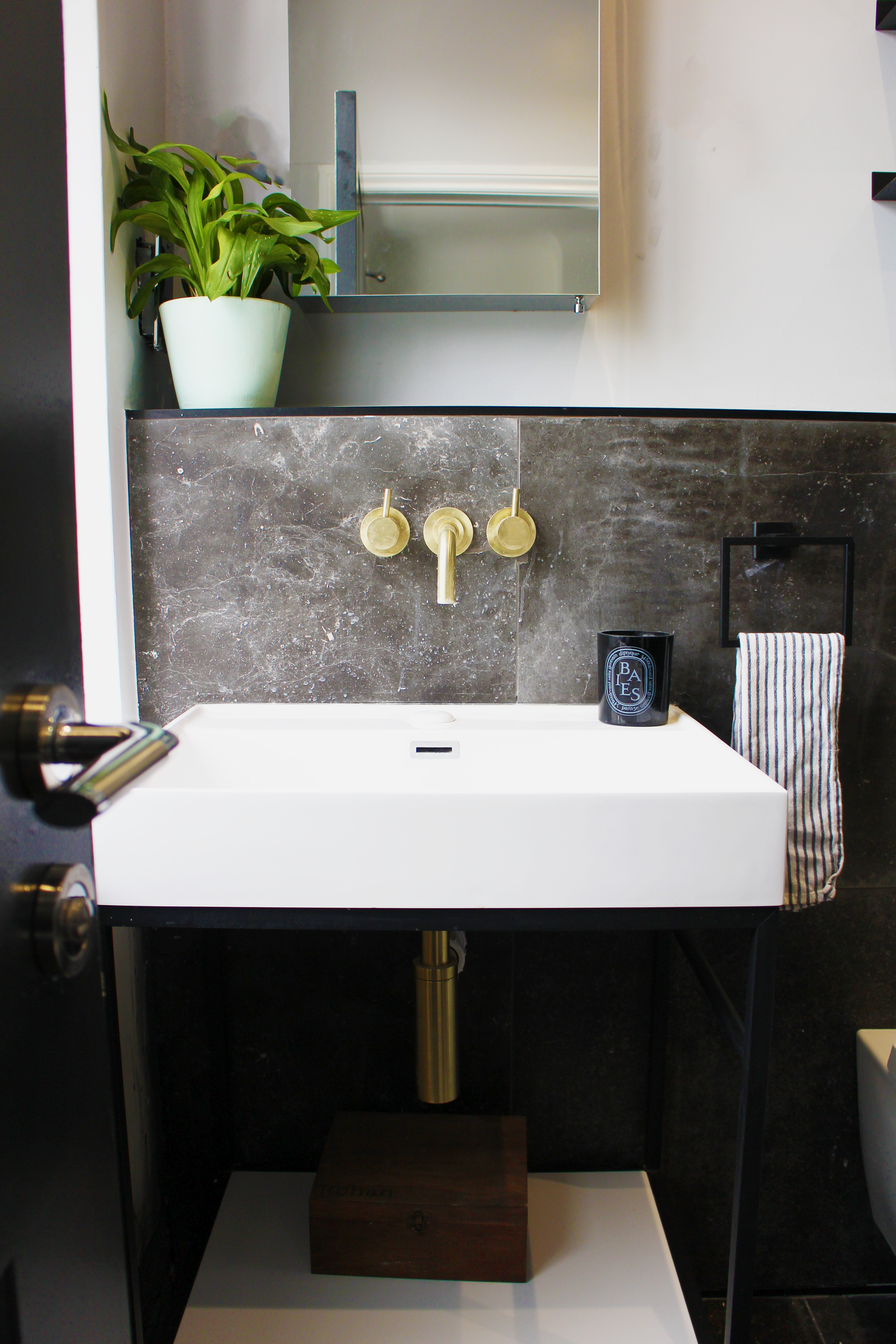 A bathroom vanity with a white rectangular sink, brass fixtures, a black mirror, a green potted plant, a black cup, and a striped towel.
