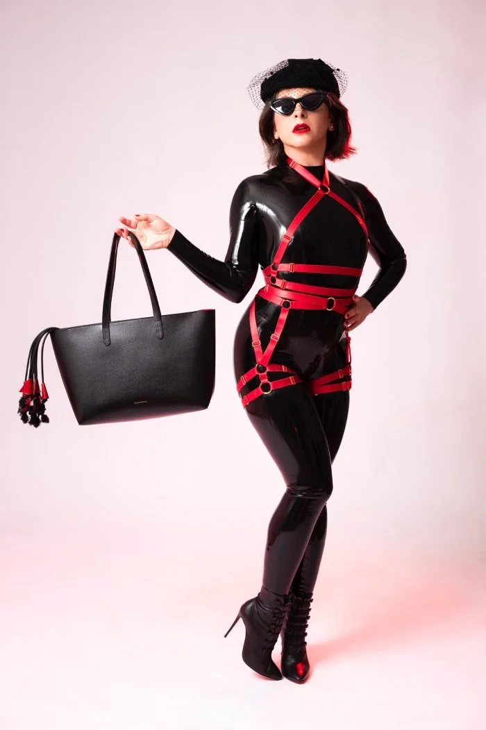 NYC Dominatrix in latex catsuit. Beautiful Mistress with perfect butt, red lips, leather and nylon. Falls from a flogger or whip stick out of her leather luxury handbag
