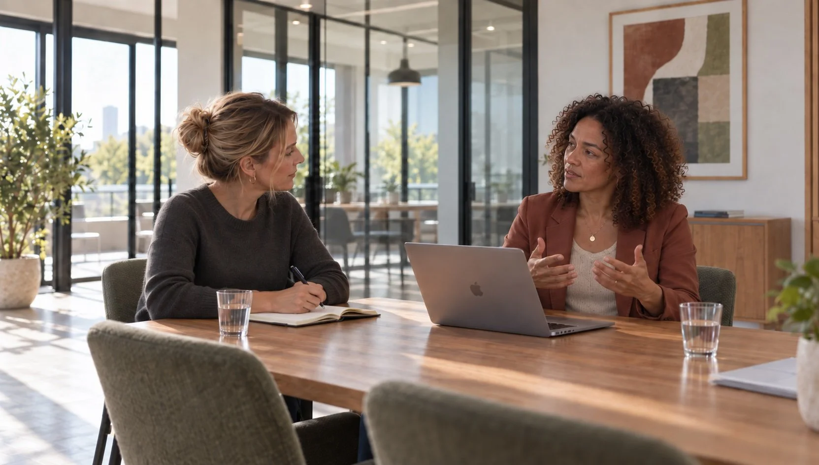 Two women having a conversation at a desk with notebooks, a laptop, a glass of water, and a potted plant in the background.