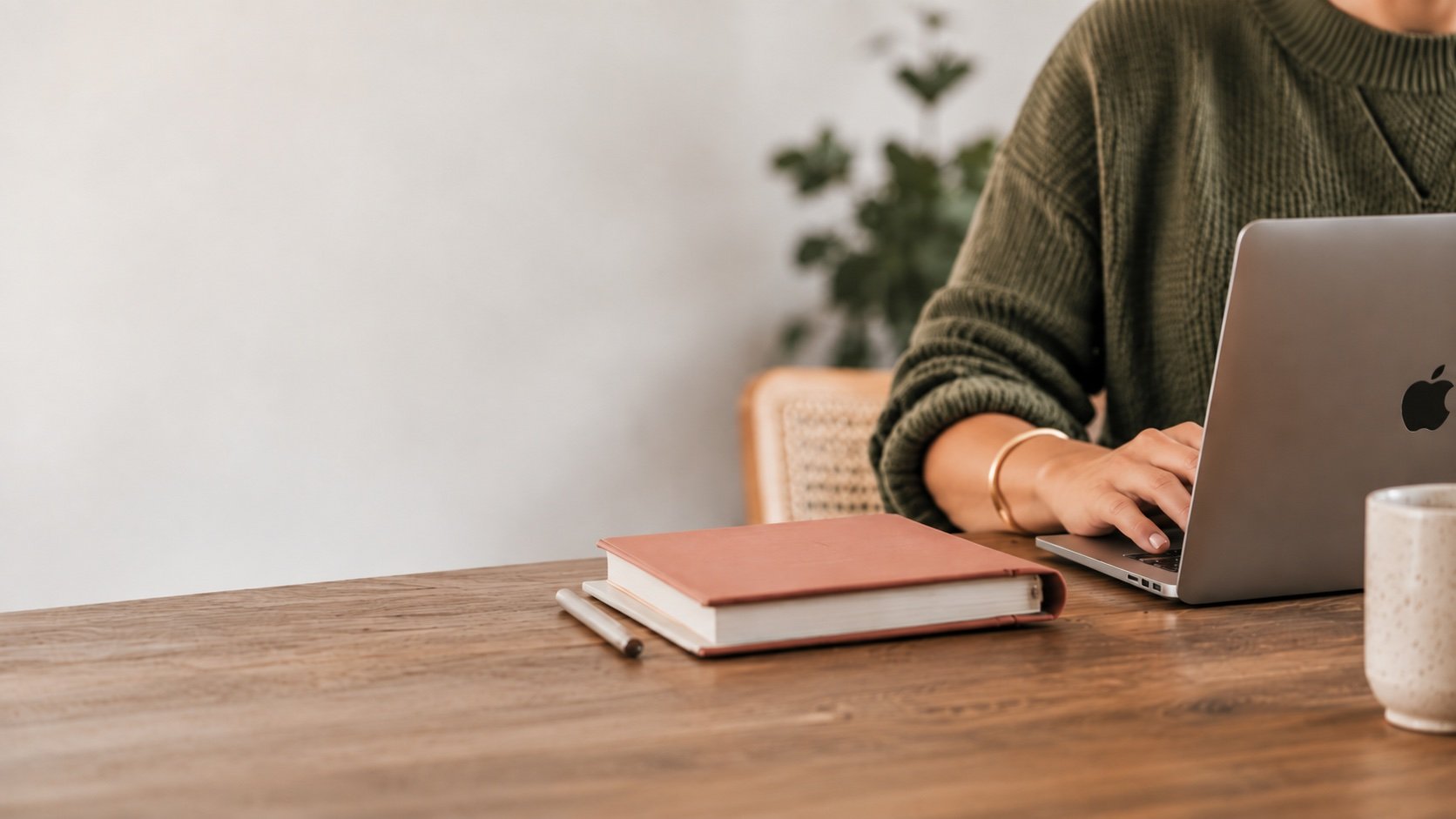 Person wearing a green sweater working on a MacBook at a wooden table with a closed pink notebook and a pen, with a plant and a white cup in the background.