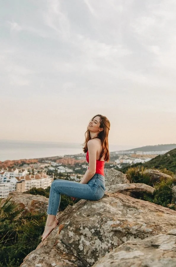 Woman sitting on a rock with a cityscape and ocean in the background, wearing a red top and jeans, smiling.