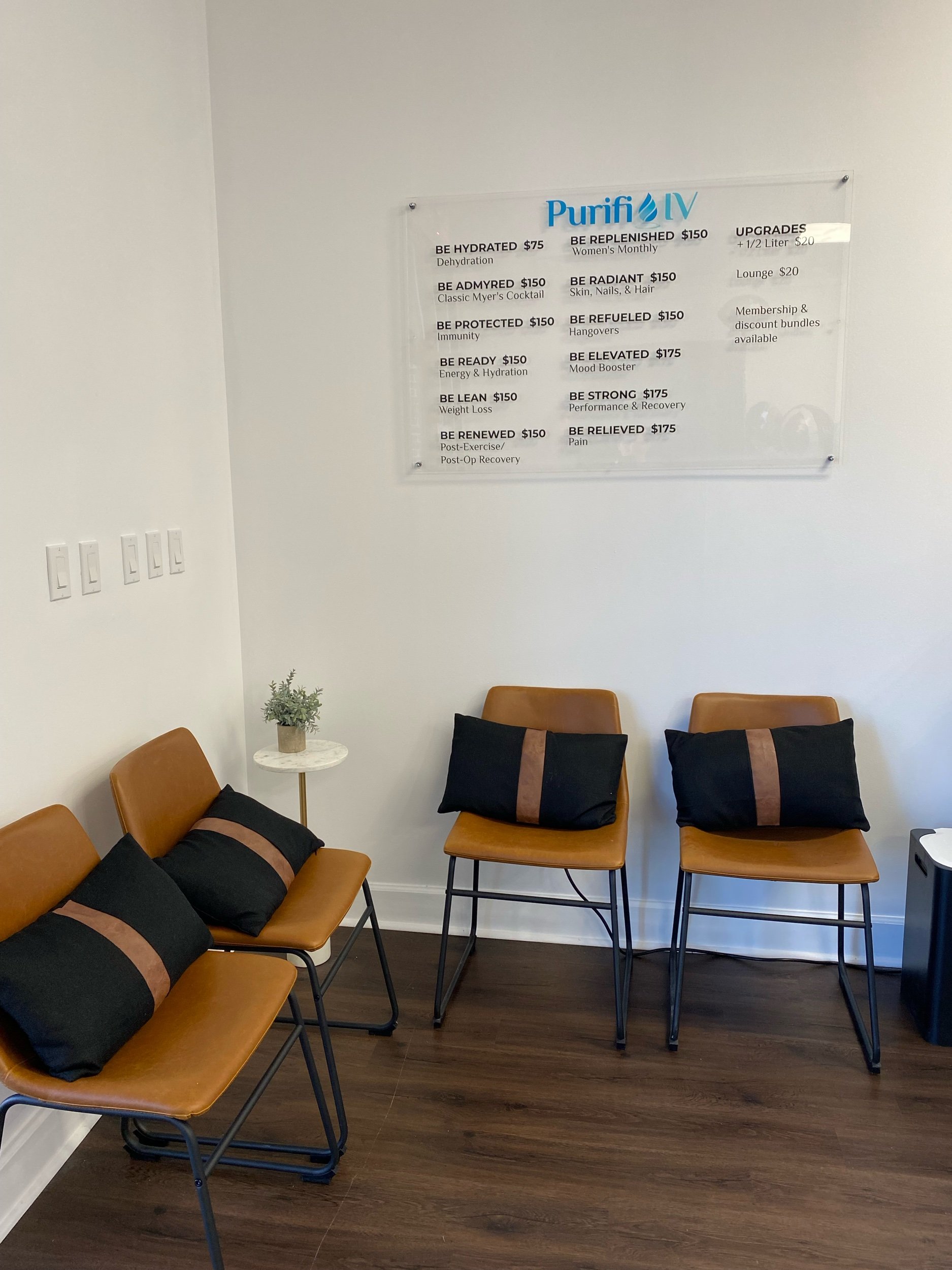 A waiting room with brown chairs, black pillows, and a small table with a plant. A sign on the wall lists various IV hydration treatments with prices.