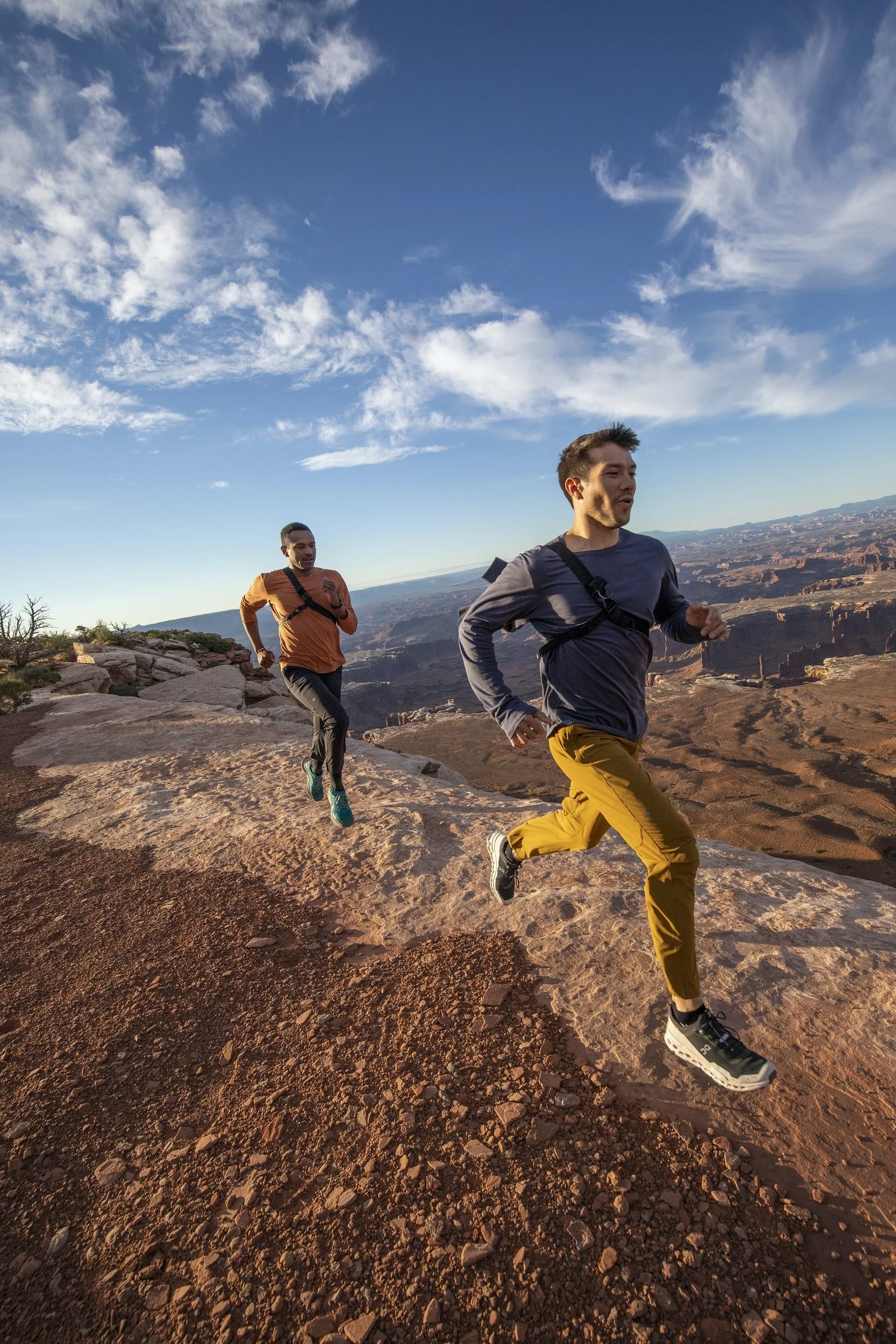 Two men trail running on a rocky path with a canyon landscape in the background under a blue sky with clouds.