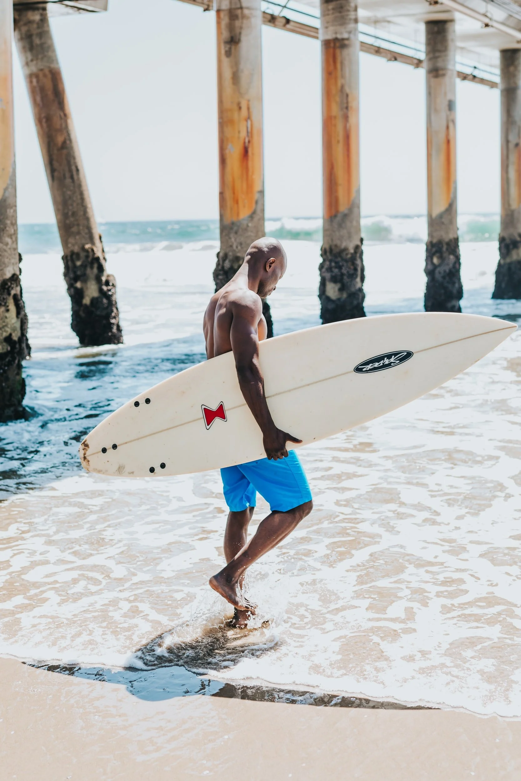 Man in blue shorts carrying a surfboard near a pier with ocean waves in the background.