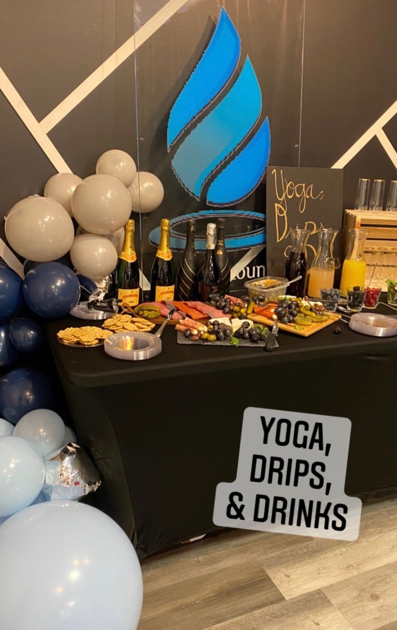 Event setup with balloons, champagne bottles, charcuterie board, and signage reading "Yoga, Drips, & Drinks" on a black table.