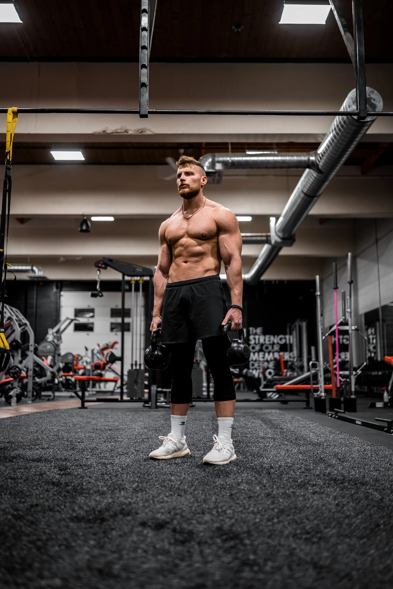 Man holding kettlebells in a gym, wearing shorts and sneakers, surrounded by gym equipment.