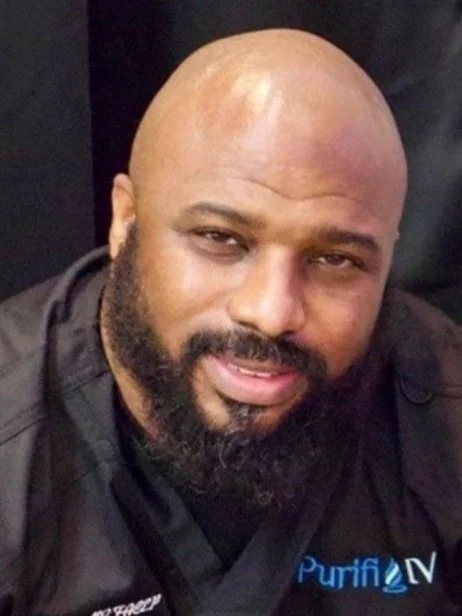 Close-up of a bald man with a beard wearing a black jacket with a logo that says 'PurifLN'.