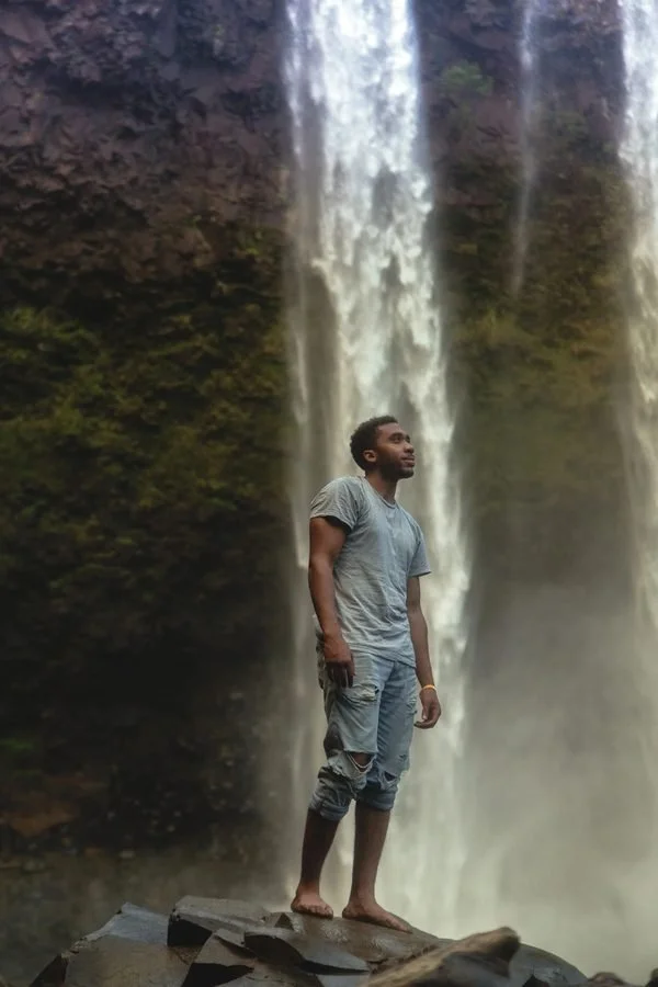Man standing barefoot on rocks in front of a waterfall.
