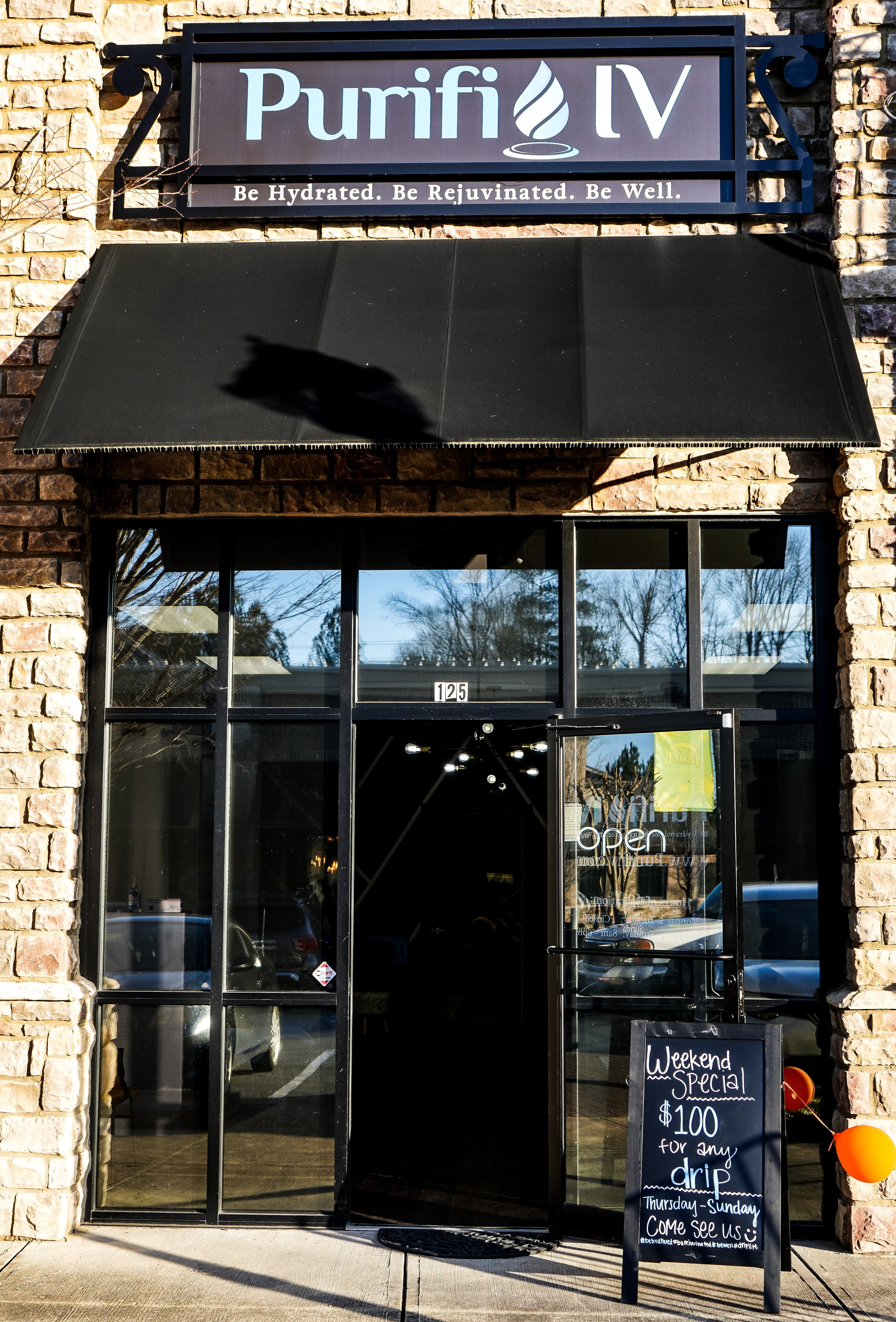 Exterior of a building with a sign reading 'Purifi IV' above a black awning, displaying a slogan: 'Be Hydrated. Be Rejuvenated. Be Well.' An entrance with glass doors shows a business open sign. A sidewalk chalkboard advertises a weekend special for