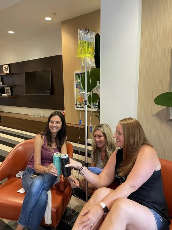 Three women sitting in a lounge area enjoying drinks, with one IV drip stand visible next to them.