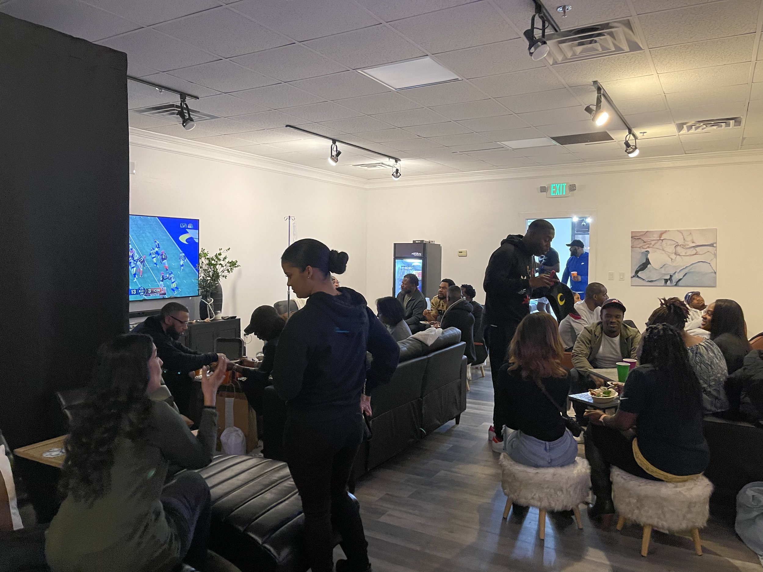 A group of people gathered in a living room watching a sports game on a large TV. The room is filled with sofas and chairs, and people are socializing, eating, and drinking. The setting is modern with overhead lights and a painting on the wall. Some