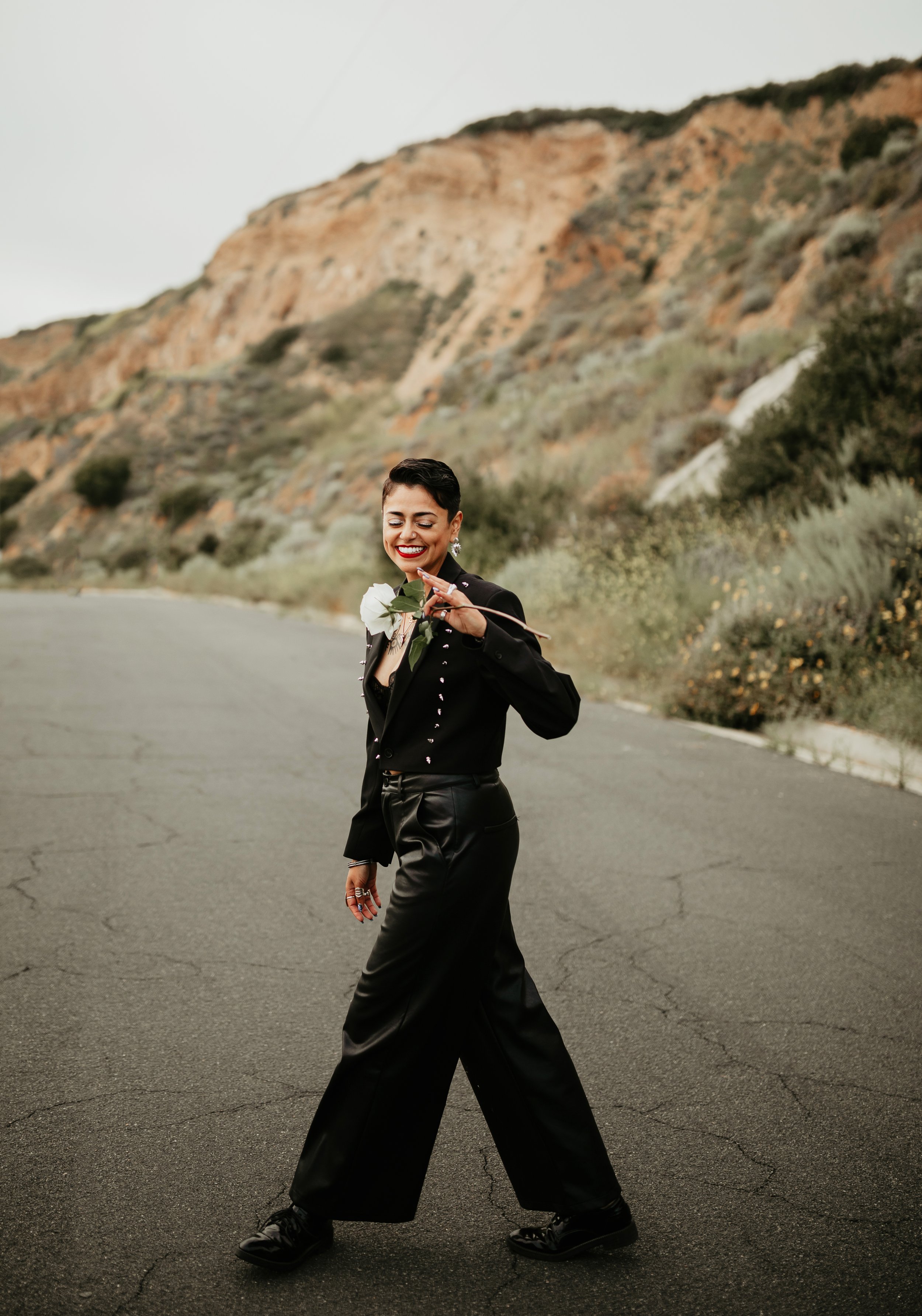 A smiling woman with short dark hair and red lipstick, dressed in a black jacket with silver embellishments and black satin wide-leg pants, standing on a cracked asphalt road with a mountain and cloudy sky in the background, holding a white flower.